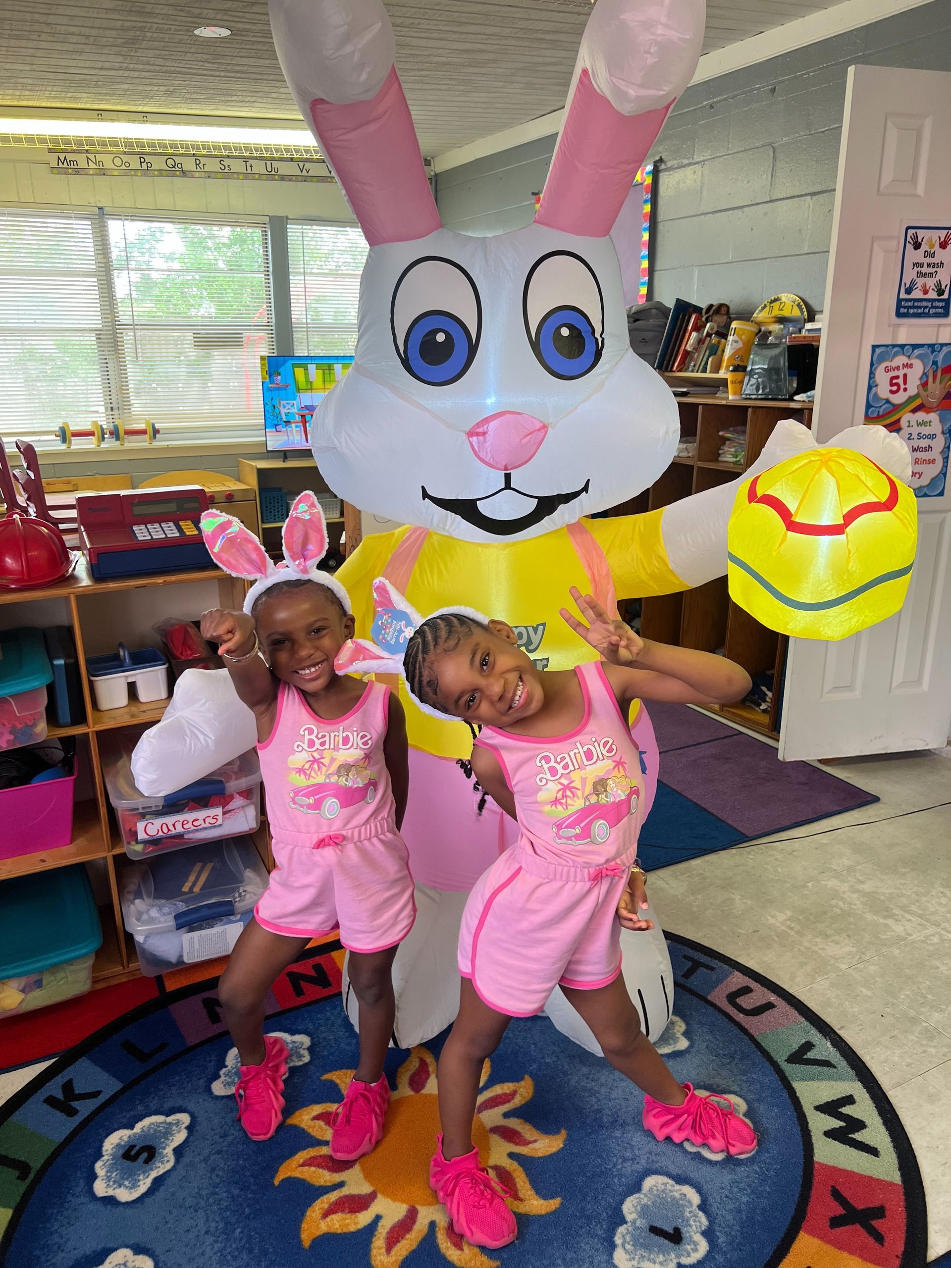 Two girls pose with an inflatable Easter bunny; both wear pink outfits and bunny ears.
