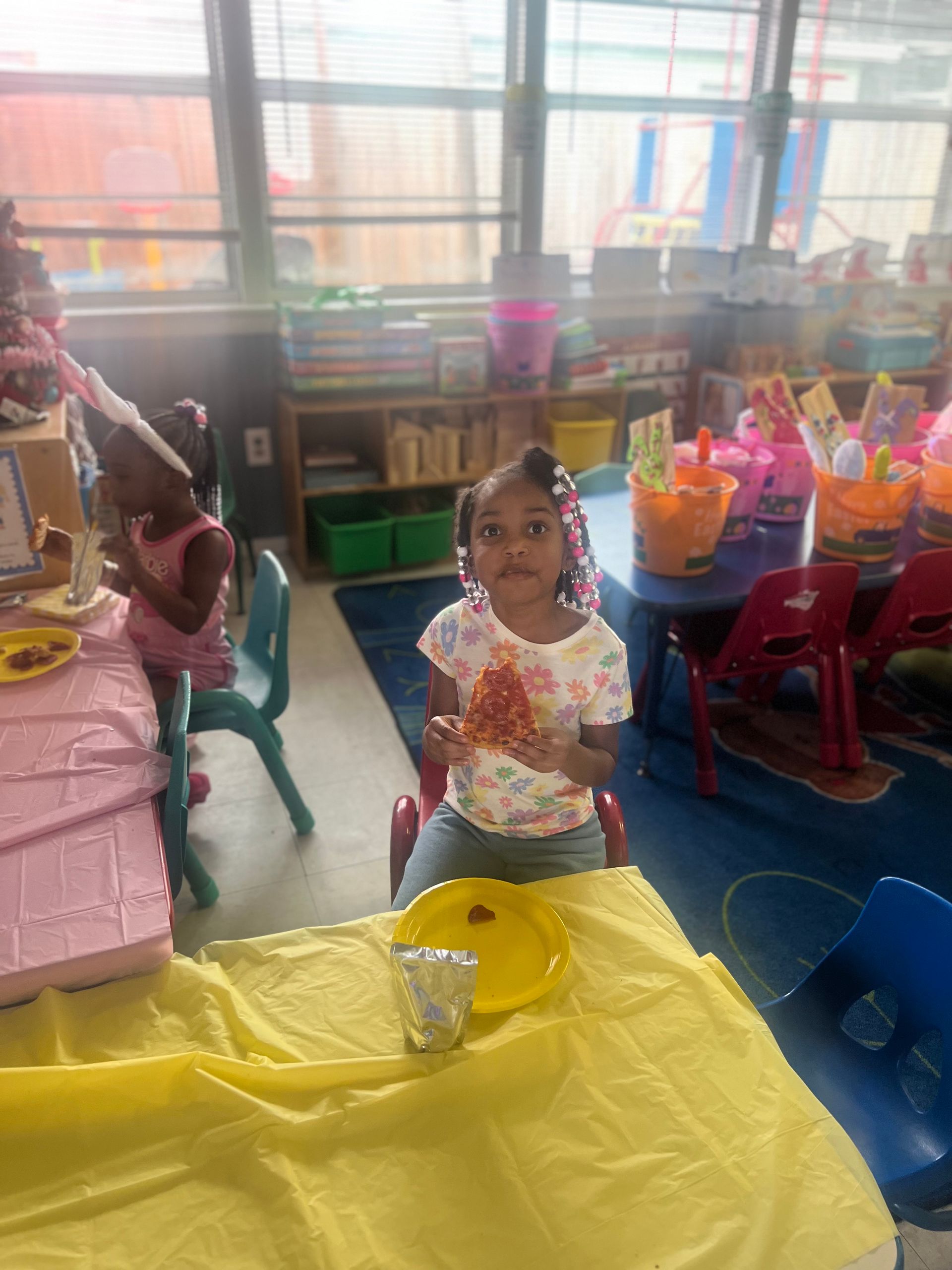 A child seated at a table, holding food. Classroom setting with other children and toys.