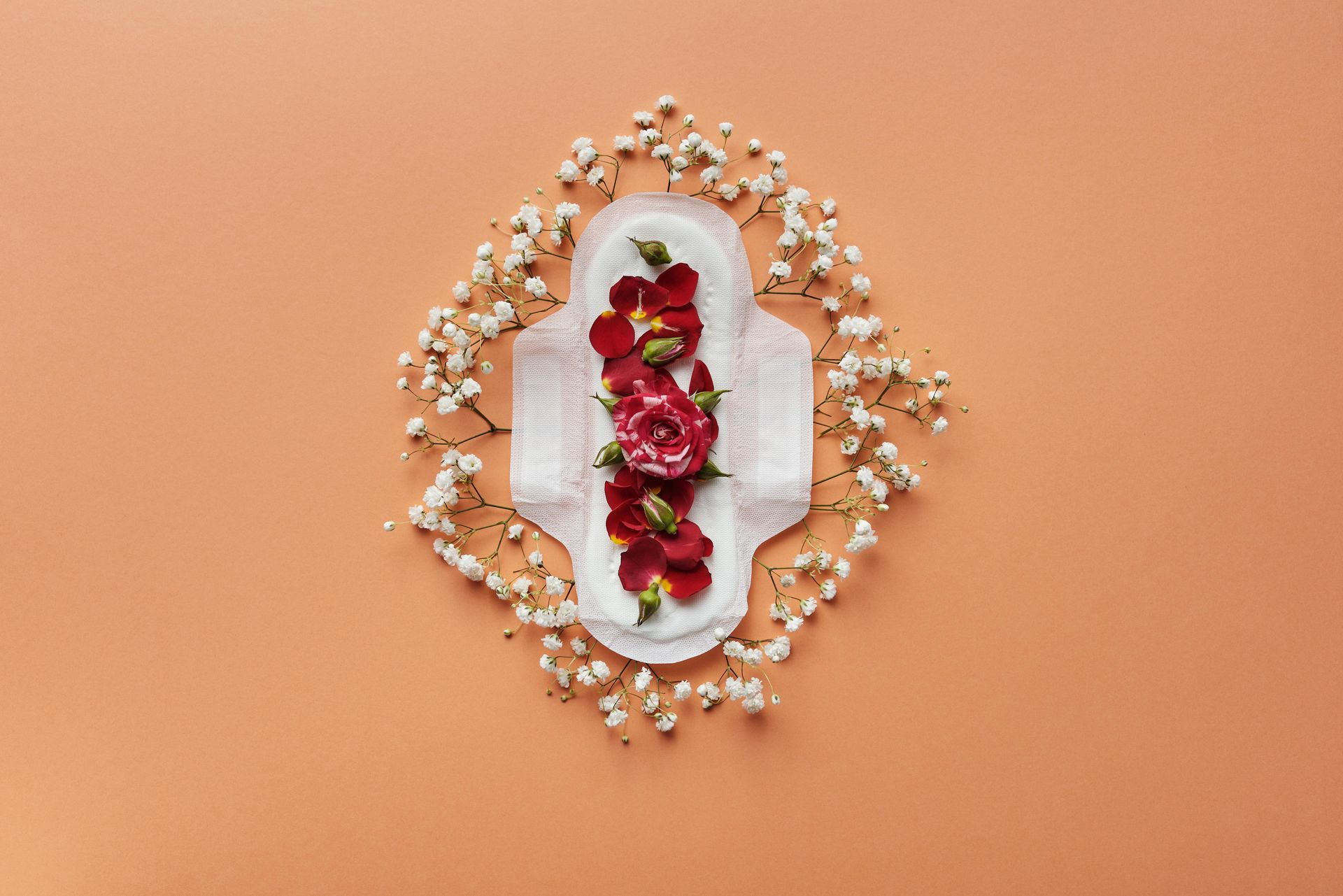 White menstrual pad with red flowers, surrounded by white flowers on an orange background.