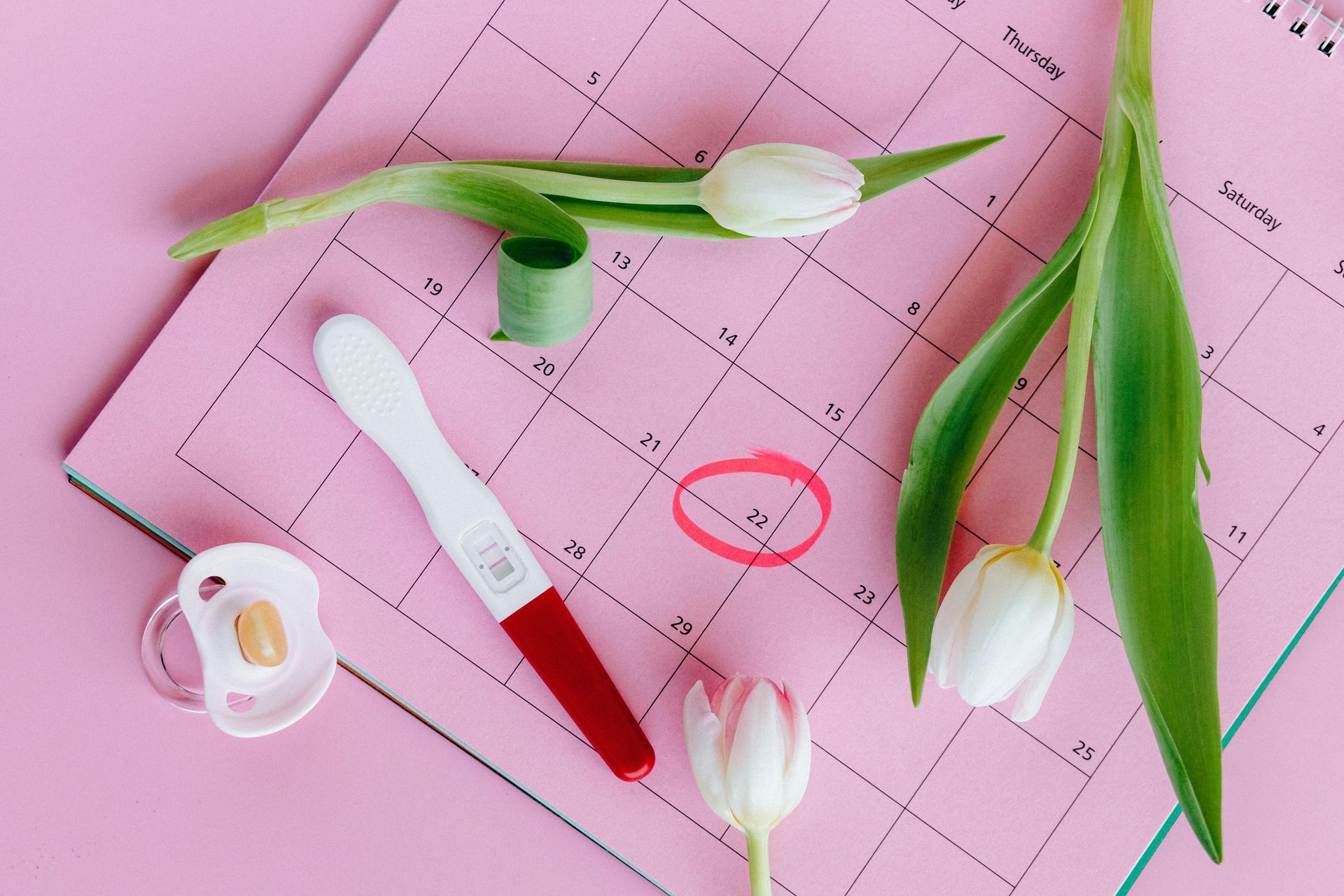 Pink calendar with circled date, pregnancy test, pacifier, and tulips.