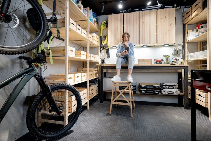A woman is sitting on a stool in a garage next to a bicycle.