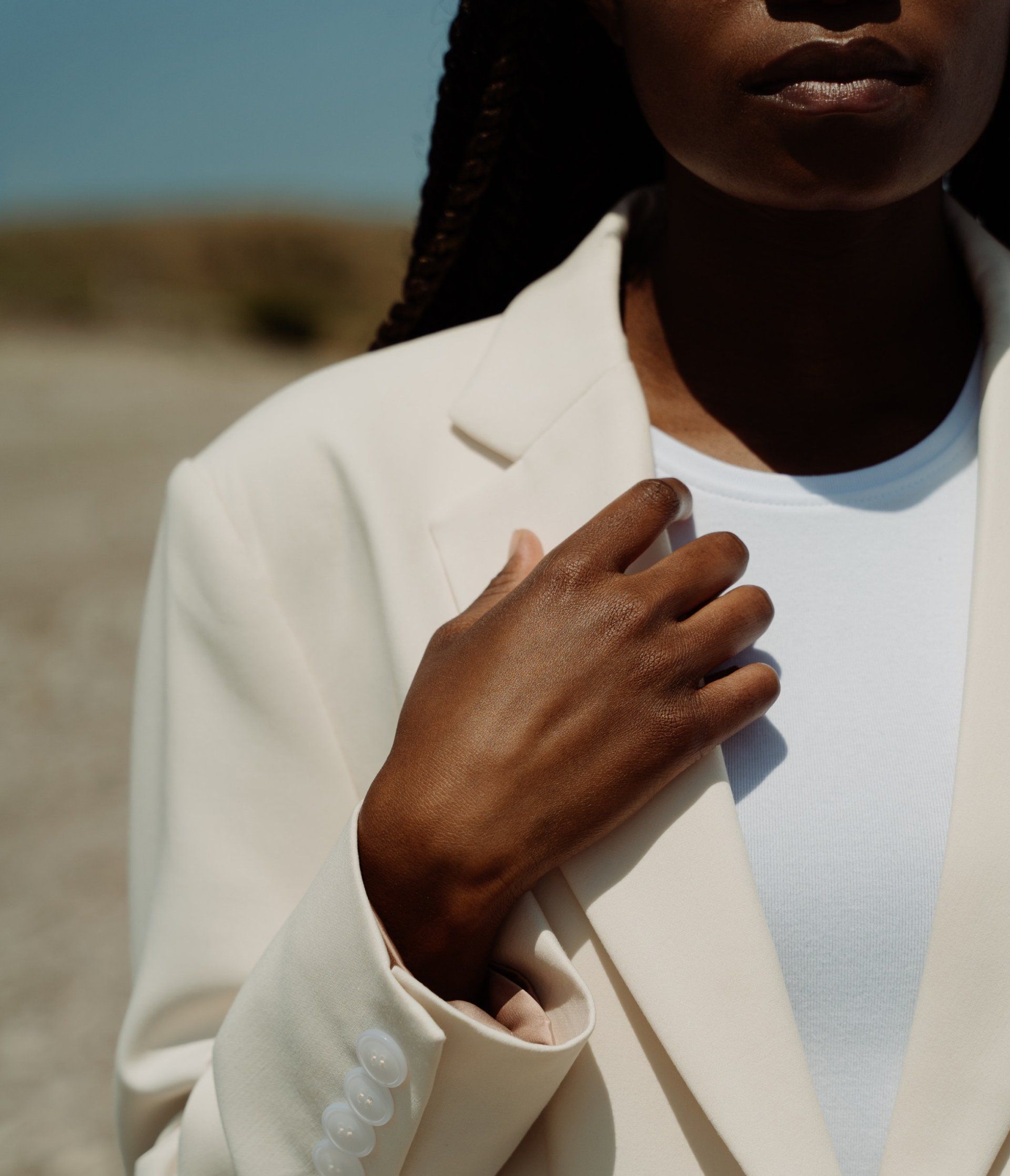 Person in a cream blazer holding the lapel, standing outdoors in sunlight.