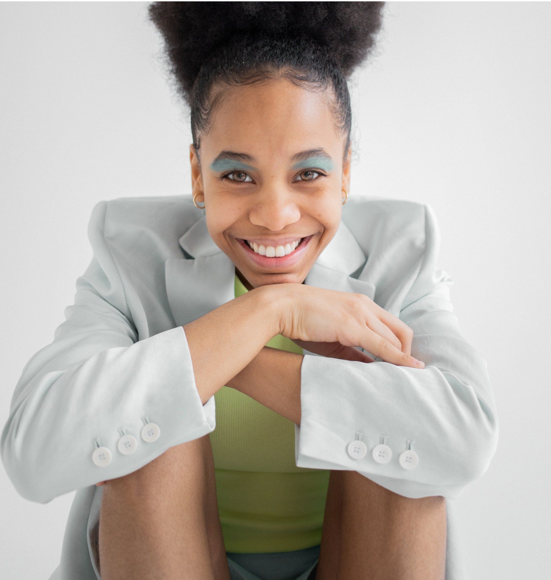 Woman in a white blazer and neon top sitting on a chair, holding a glass, against a plain gray background