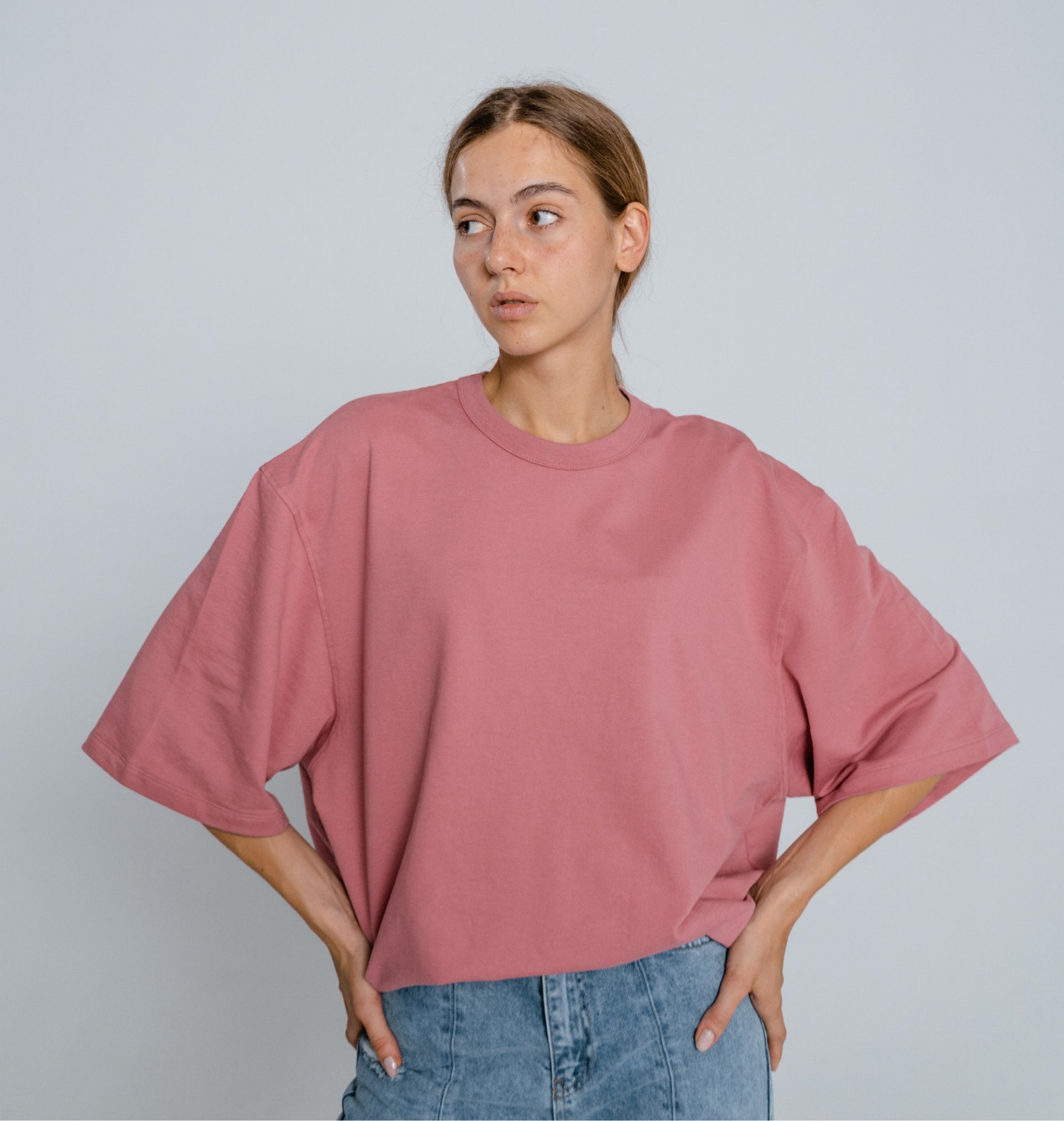 Person in a loose pink T-shirt and blue jeans standing with hands on hips against a gray background.