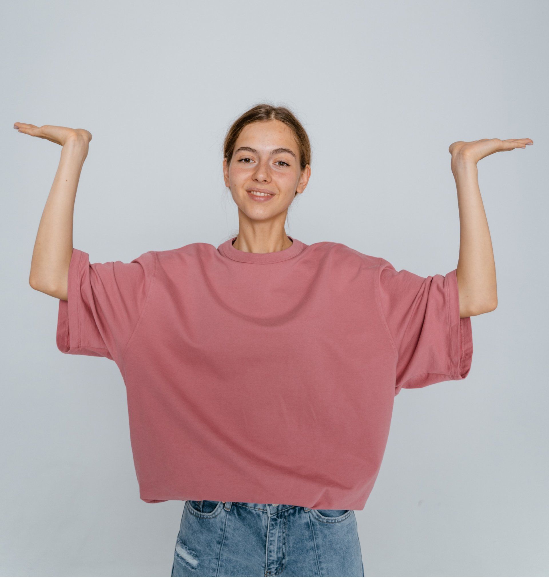 Person in a loose pink T-shirt and blue jeans standing with hands on hips against a gray background.