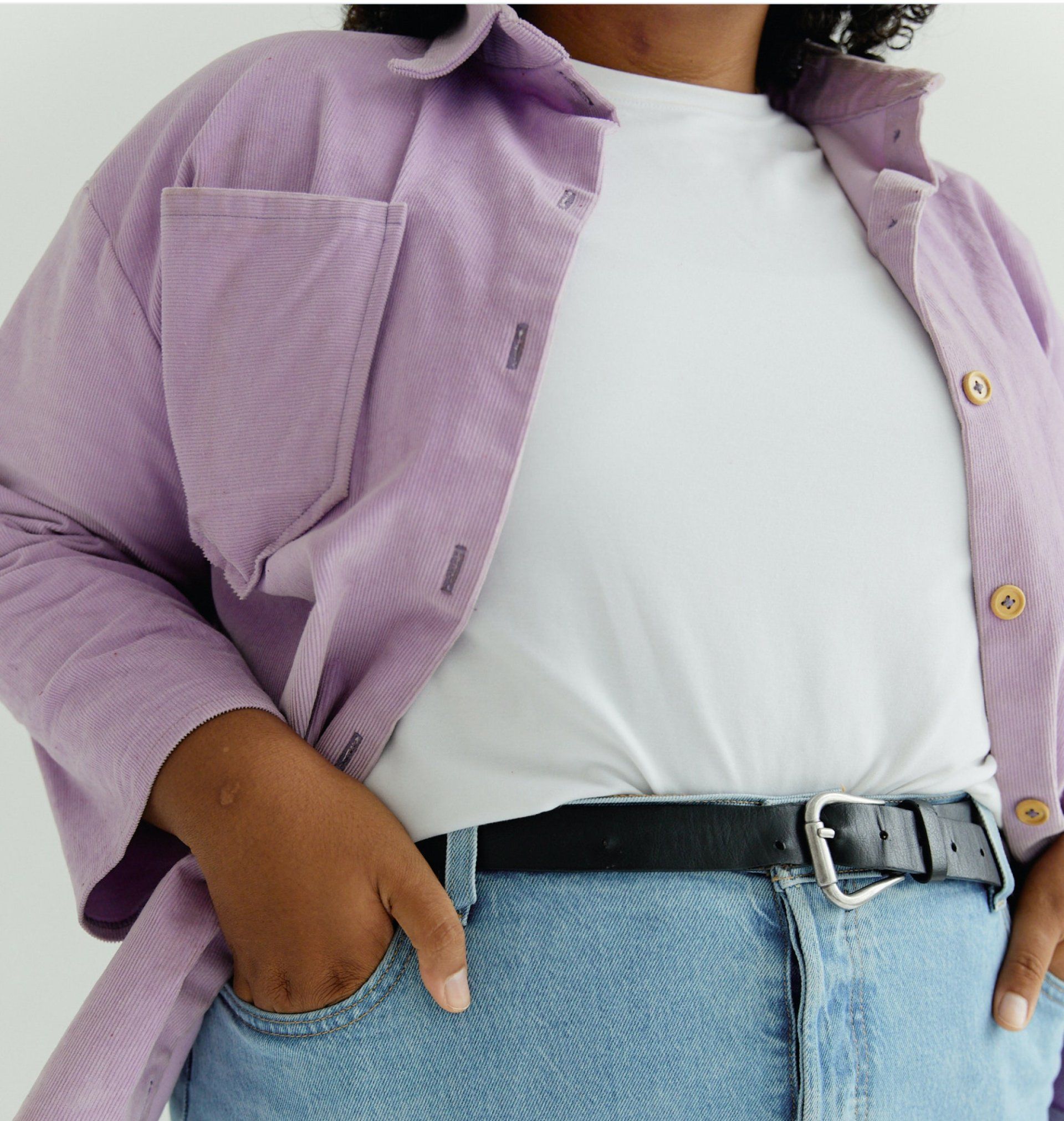 Person wearing a lavender shirt and jeans, adjusting a black belt against a light gray background