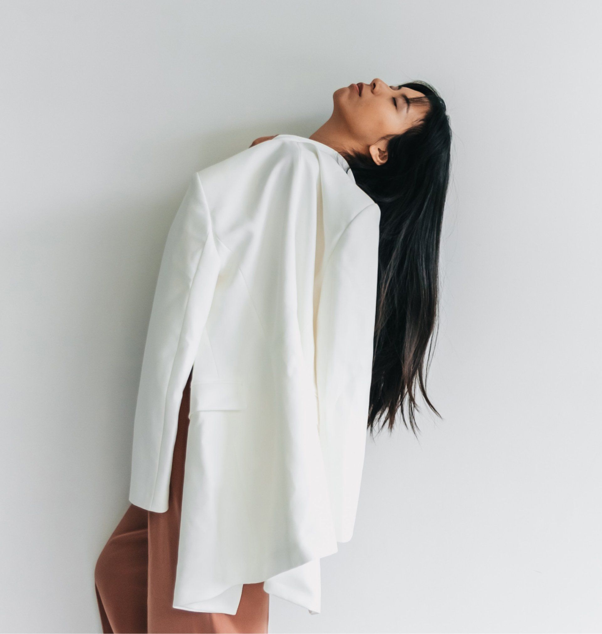 Person in a white blouse and brown pants leans back against a plain wall, looking upward.