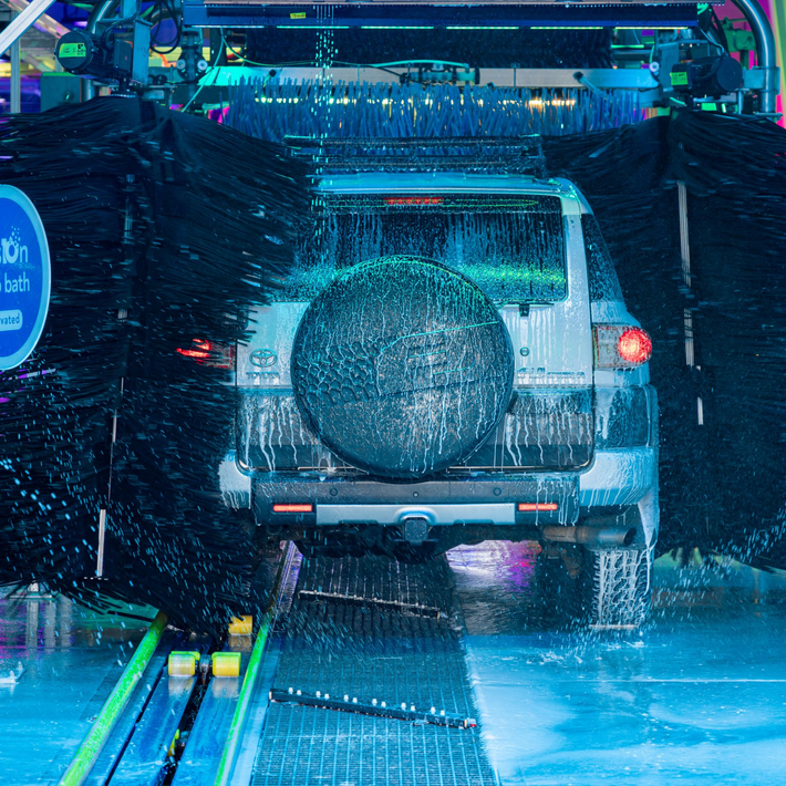 Rear view of a white truck driving through a neon-lit car wash at night, with water spraying around it