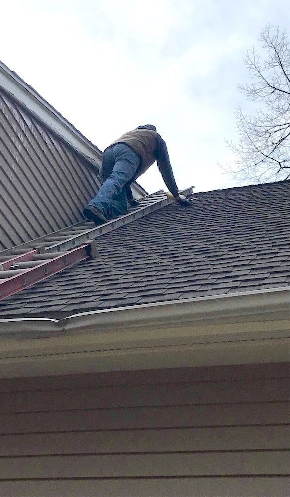 Man on roof, installing shingles. Brown roof, ladder, cloudy sky.