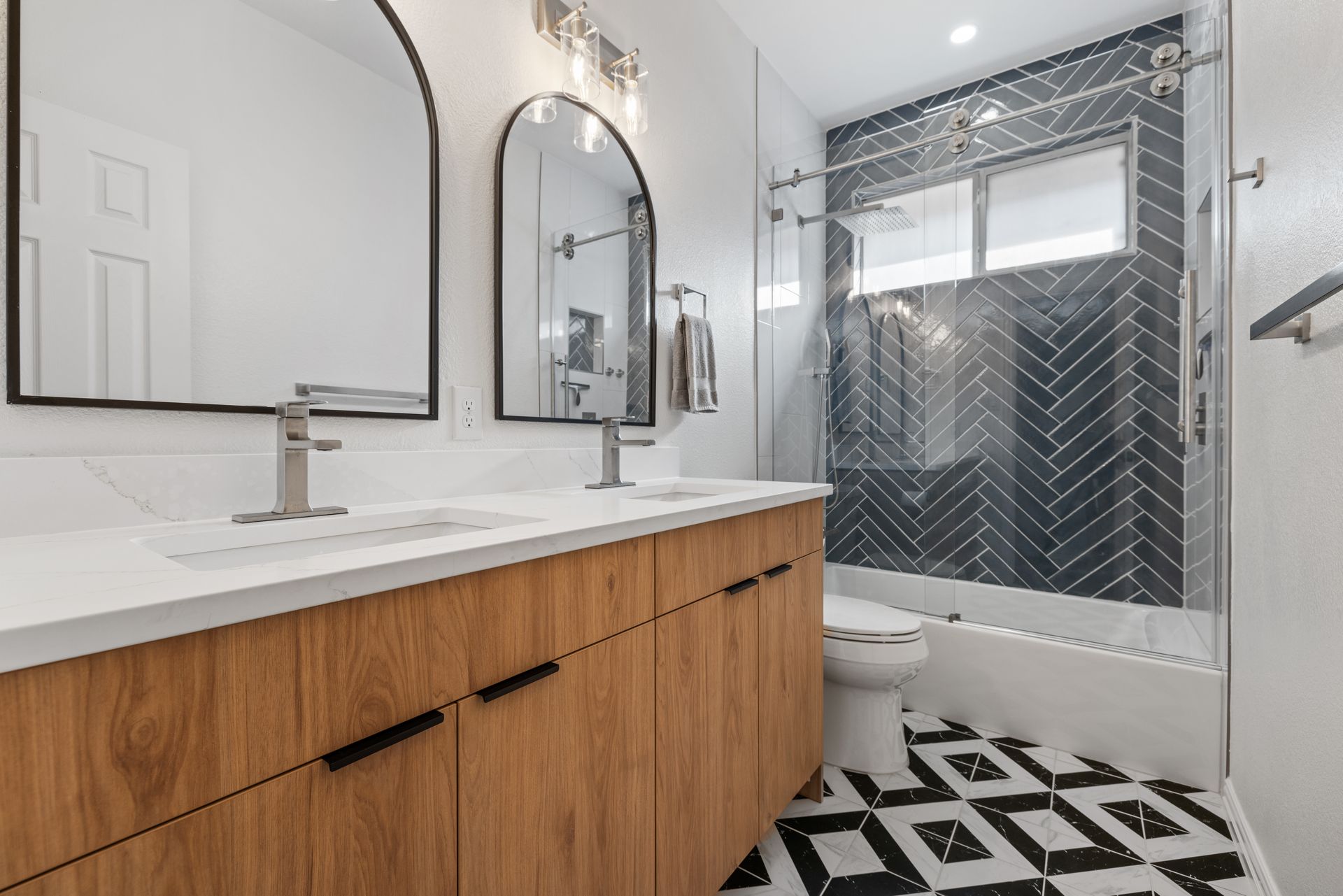 Bathroom remodel featuring wood vanity, double mirrors, black-and-white patterned tile floor, and herringbone wall tile.