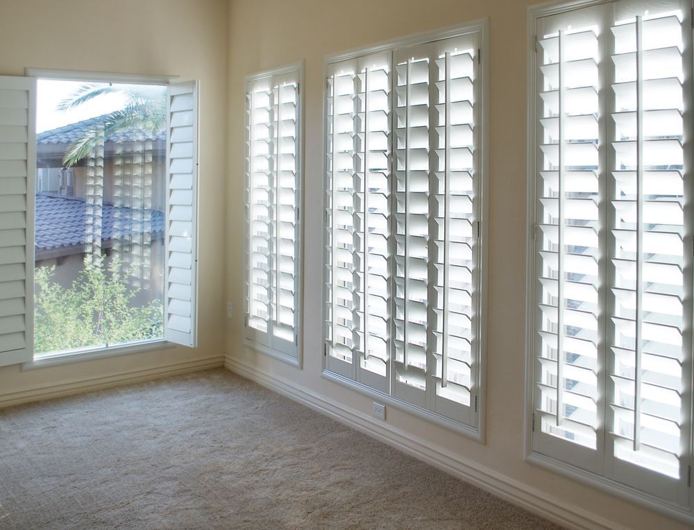A room with beige walls and carpet, featuring four windows covered with white plantation shutters.