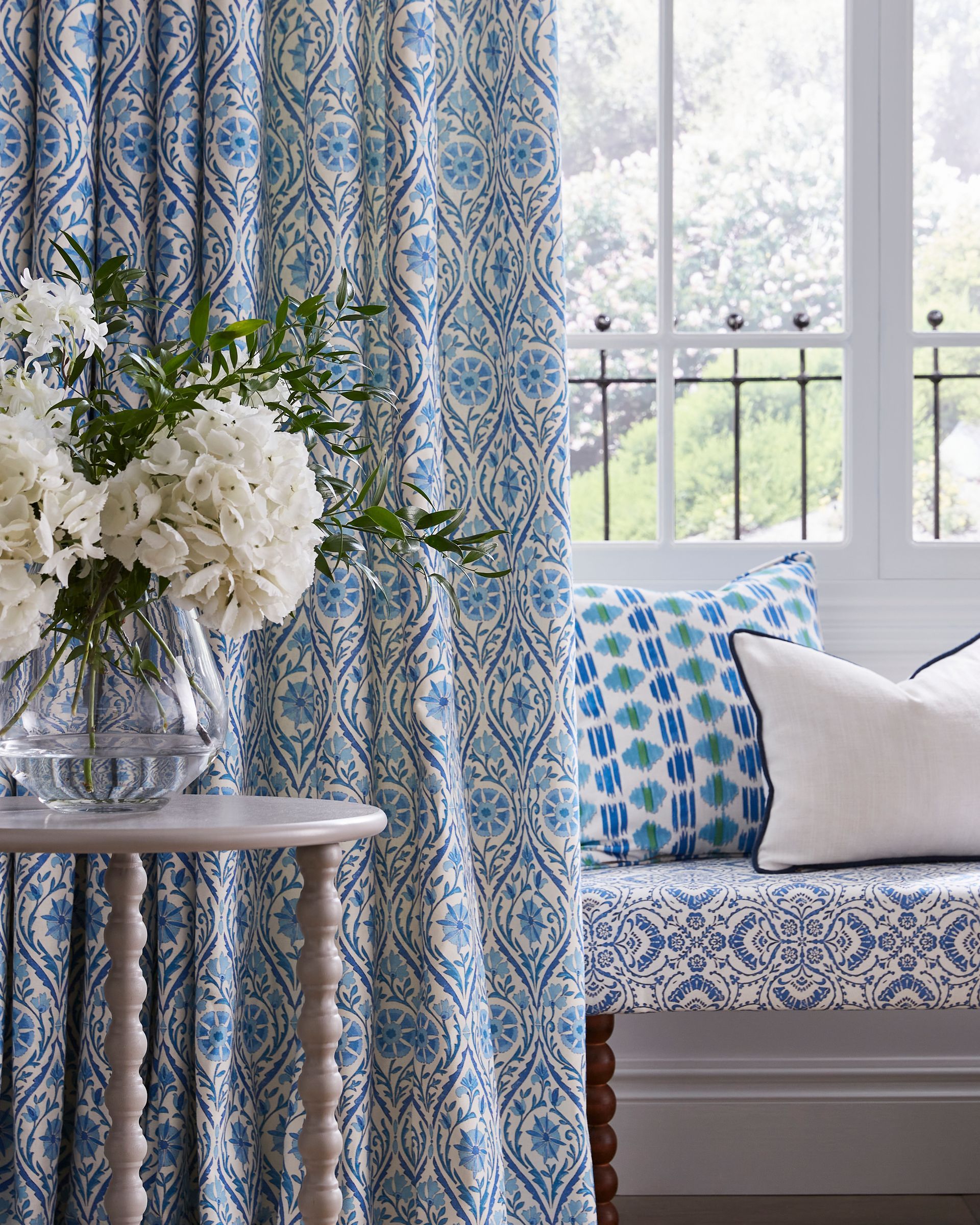Blue and white patterned curtains hang next to a window seat with matching pillows and a side table holding white flowers.