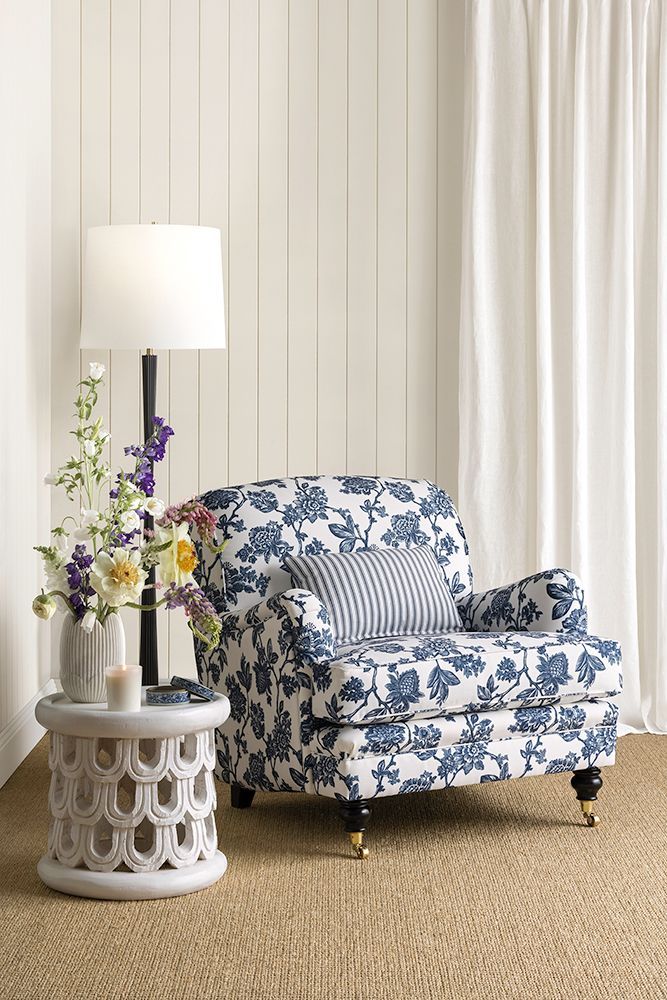 Armchair with blue floral pattern and striped pillow, next to a side table with a lamp, flowers, and a white curtain.