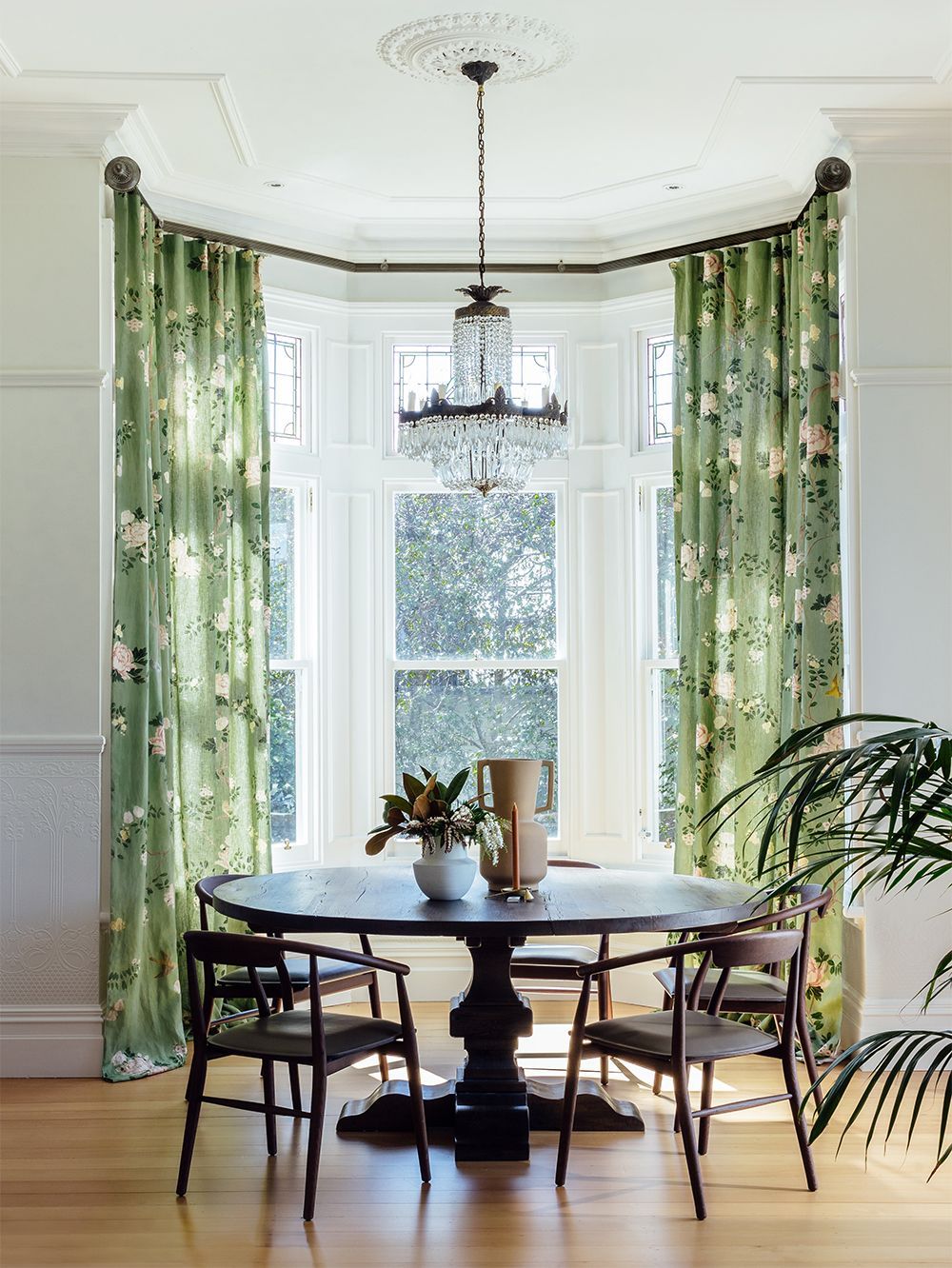 Dining room with round table, chairs, floral green curtains, chandelier, and bay windows.