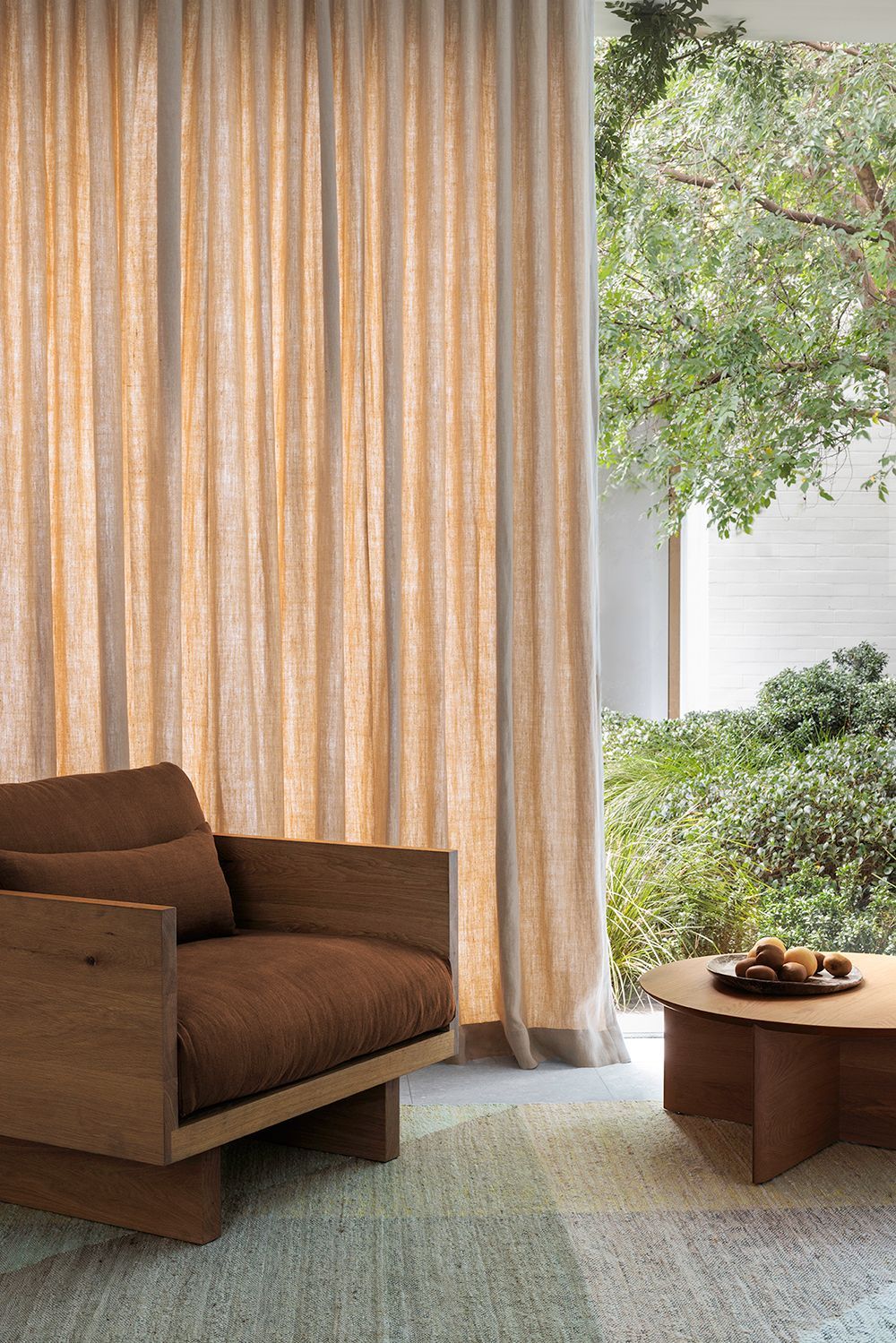 A brown armchair and low wooden coffee table sit on a textured rug before floor-to-ceiling curtains with a garden view.