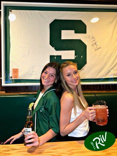 Two women standing back to back holding drinks in front of a large s.