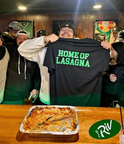 A man is holding up a shirt that says home of lasagna