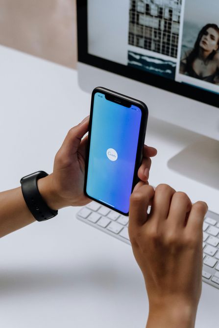 A person is holding a cell phone in front of a computer.
