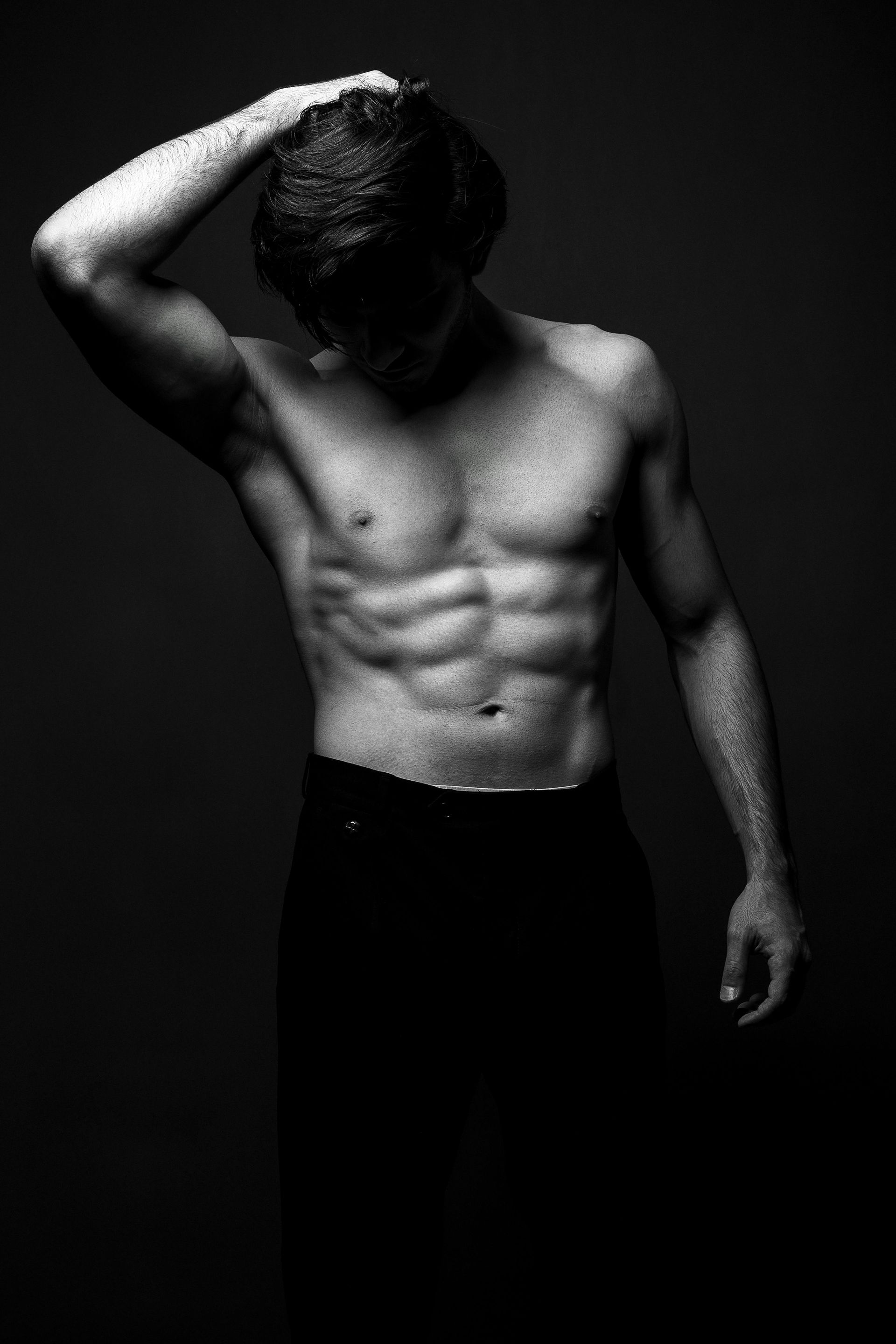 A shirtless man is standing in a black and white photo with his hand on his head.