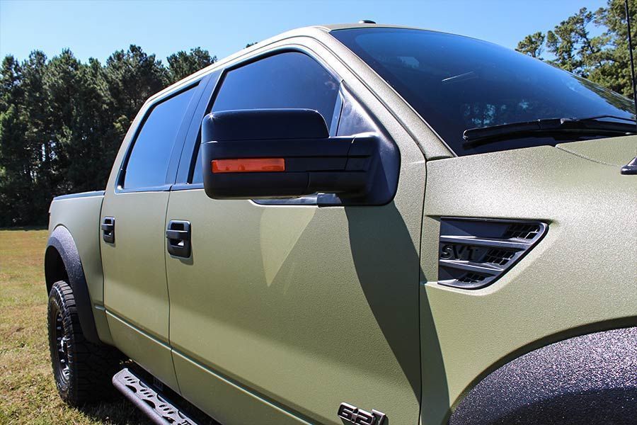Un pick-up Ford Raptor vert olive avec des garnitures noires, garé à l'extérieur par une journée ensoleillée.