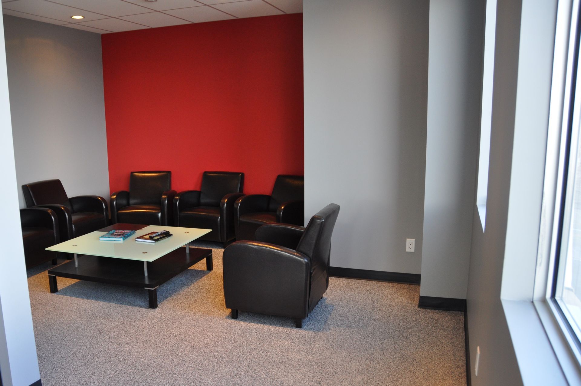 Salle d'attente avec fauteuils en cuir, table basse, mur d'accent rouge et fenêtre.