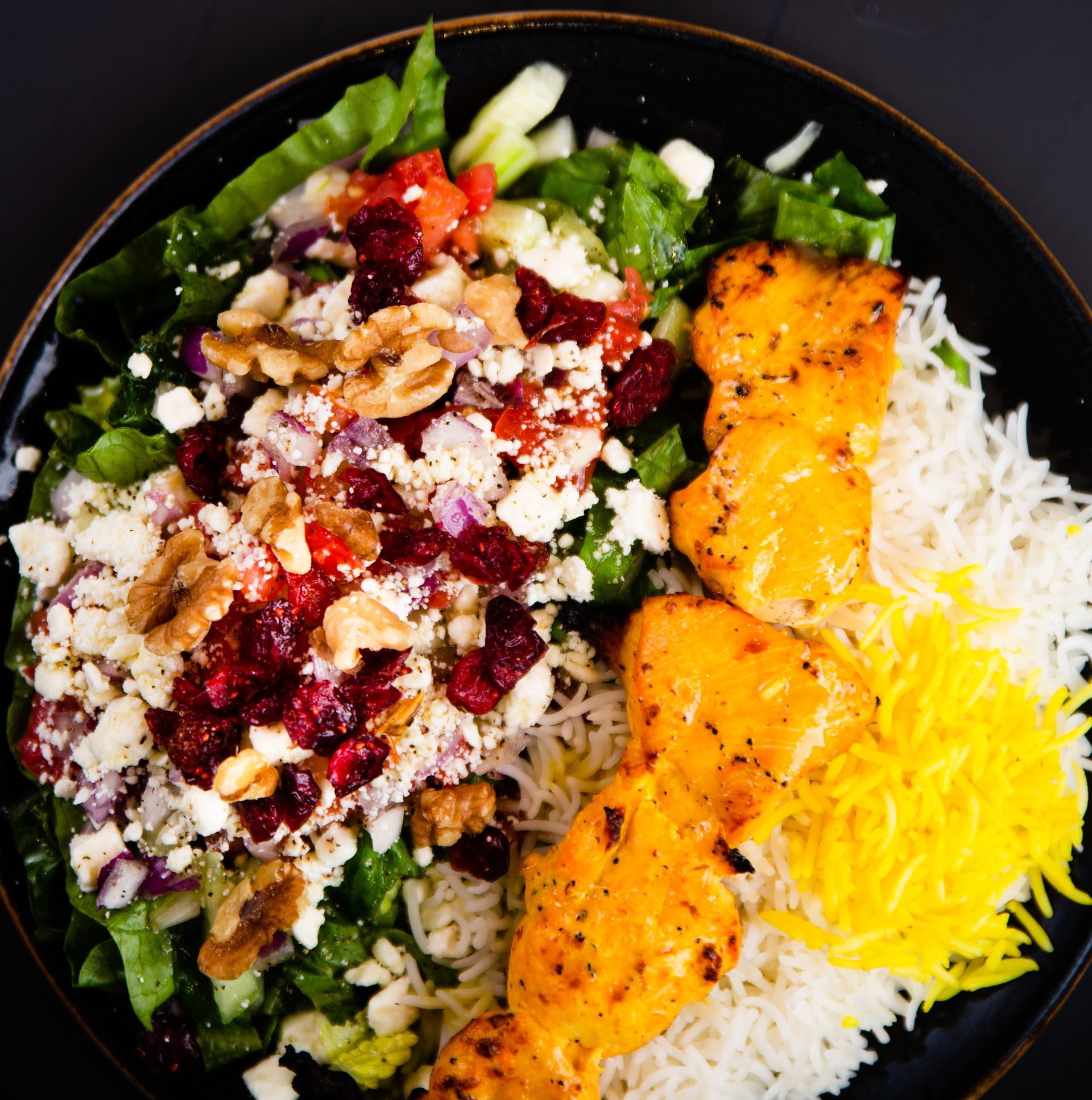 A black plate topped with rice , chicken , cranberries , and cheese.