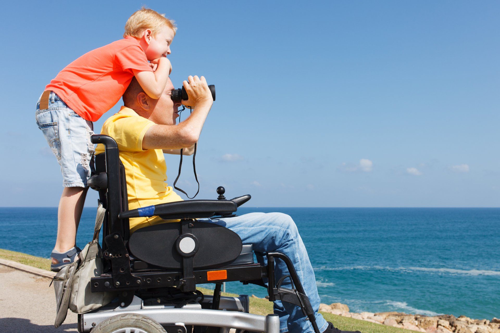 A boy is standing next to a man in a wheelchair looking through binoculars.