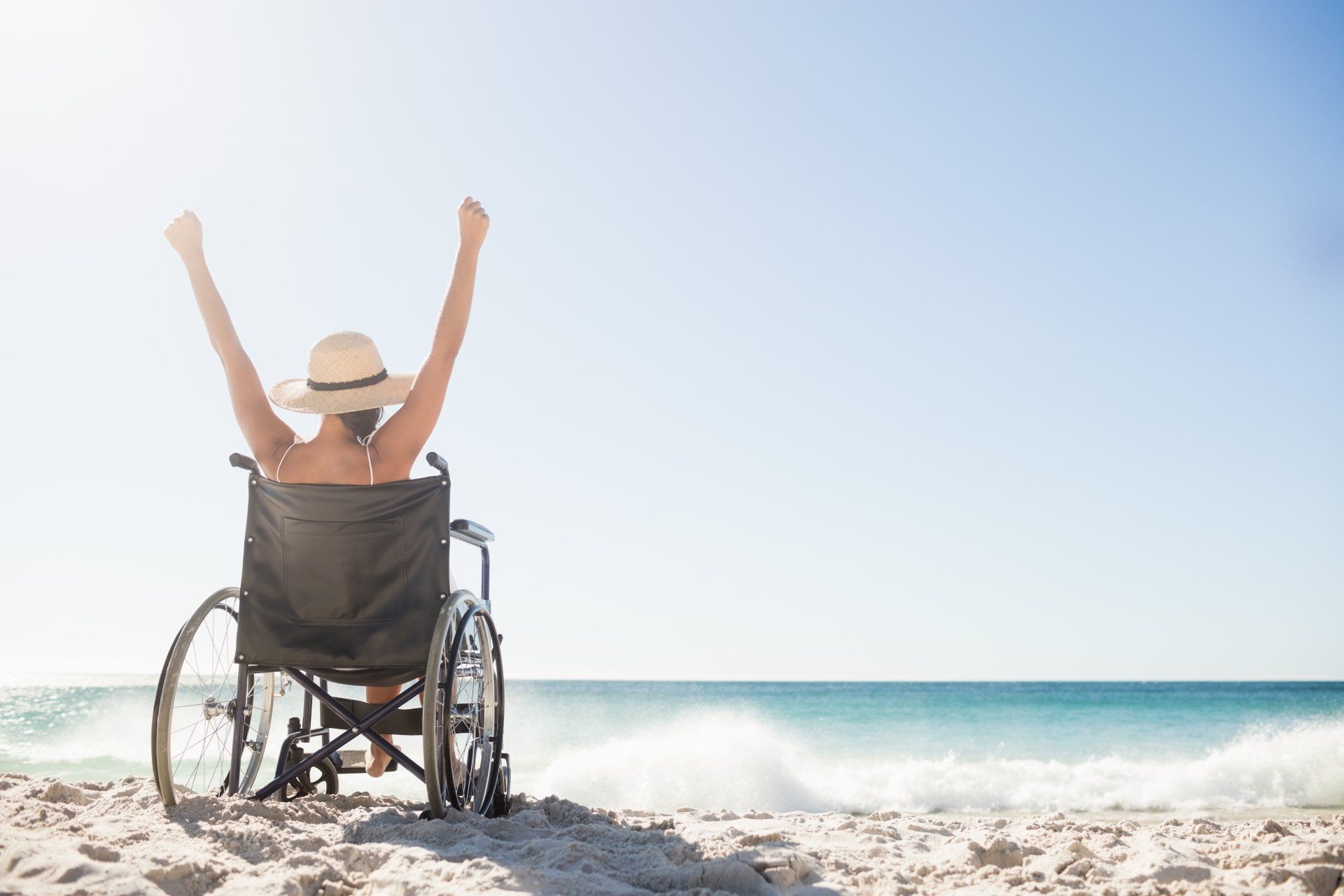 A woman in a wheelchair is sitting on the beach with her arms in the air.