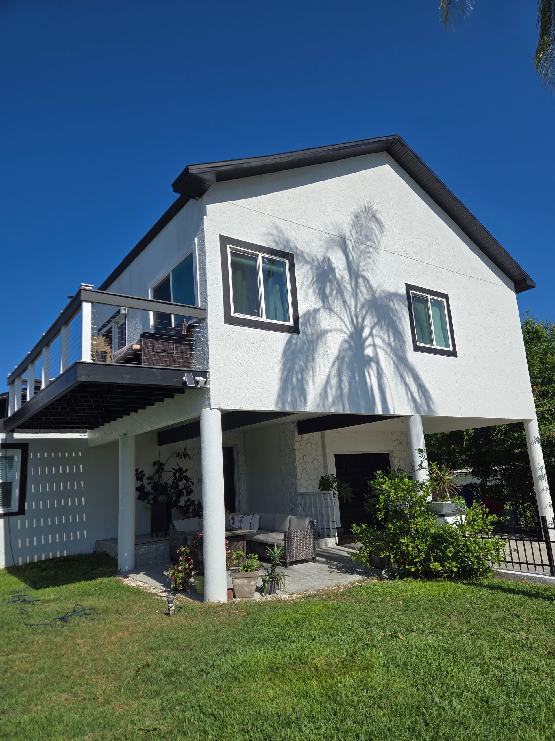 White two-story house with a black roof and balcony, set against a blue sky, with a grassy lawn.