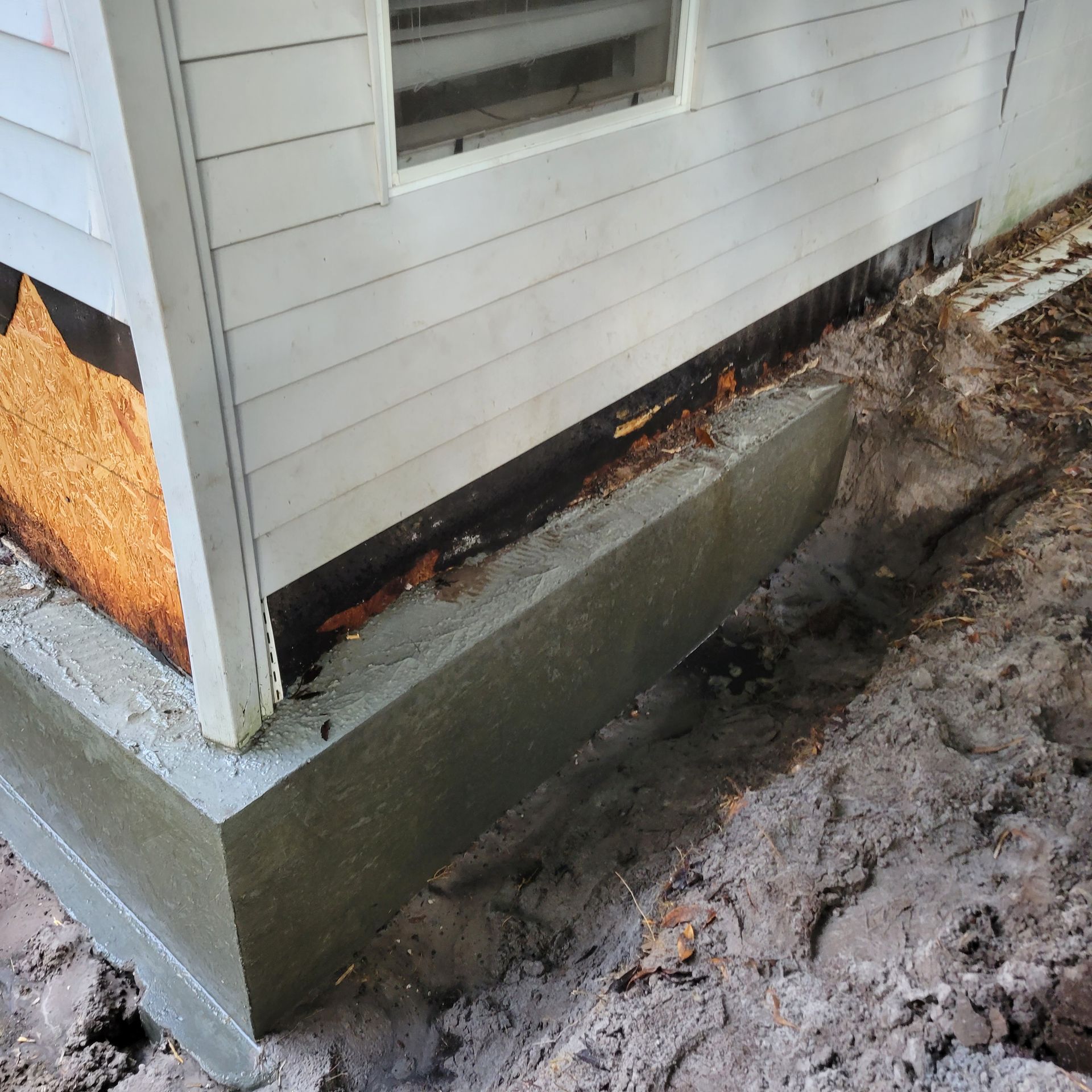 Exterior view of a house foundation, showing freshly poured concrete and surrounding soil.