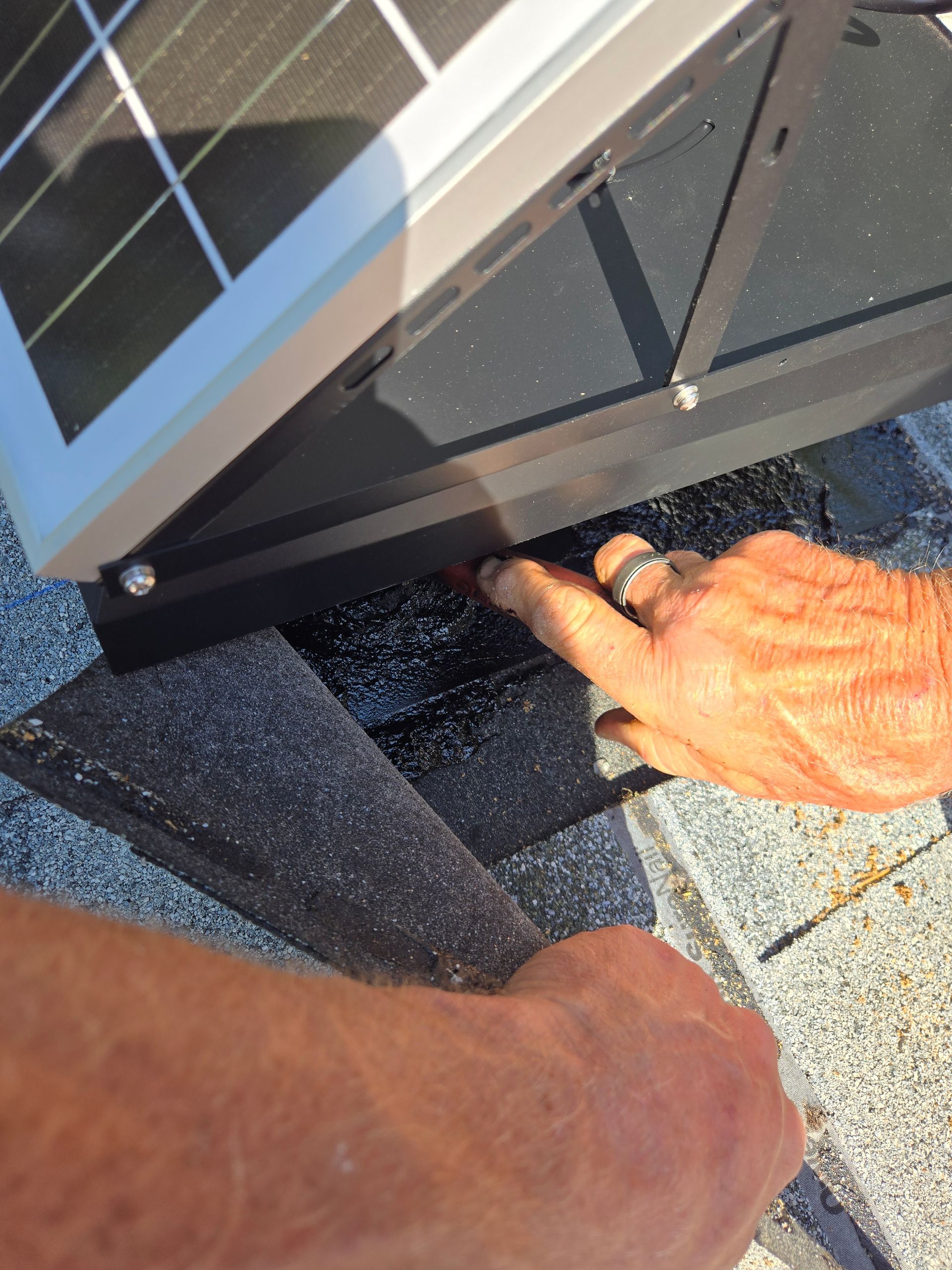 Hands near a solar panel mounted on a shingle roof, possibly sealing around the base.