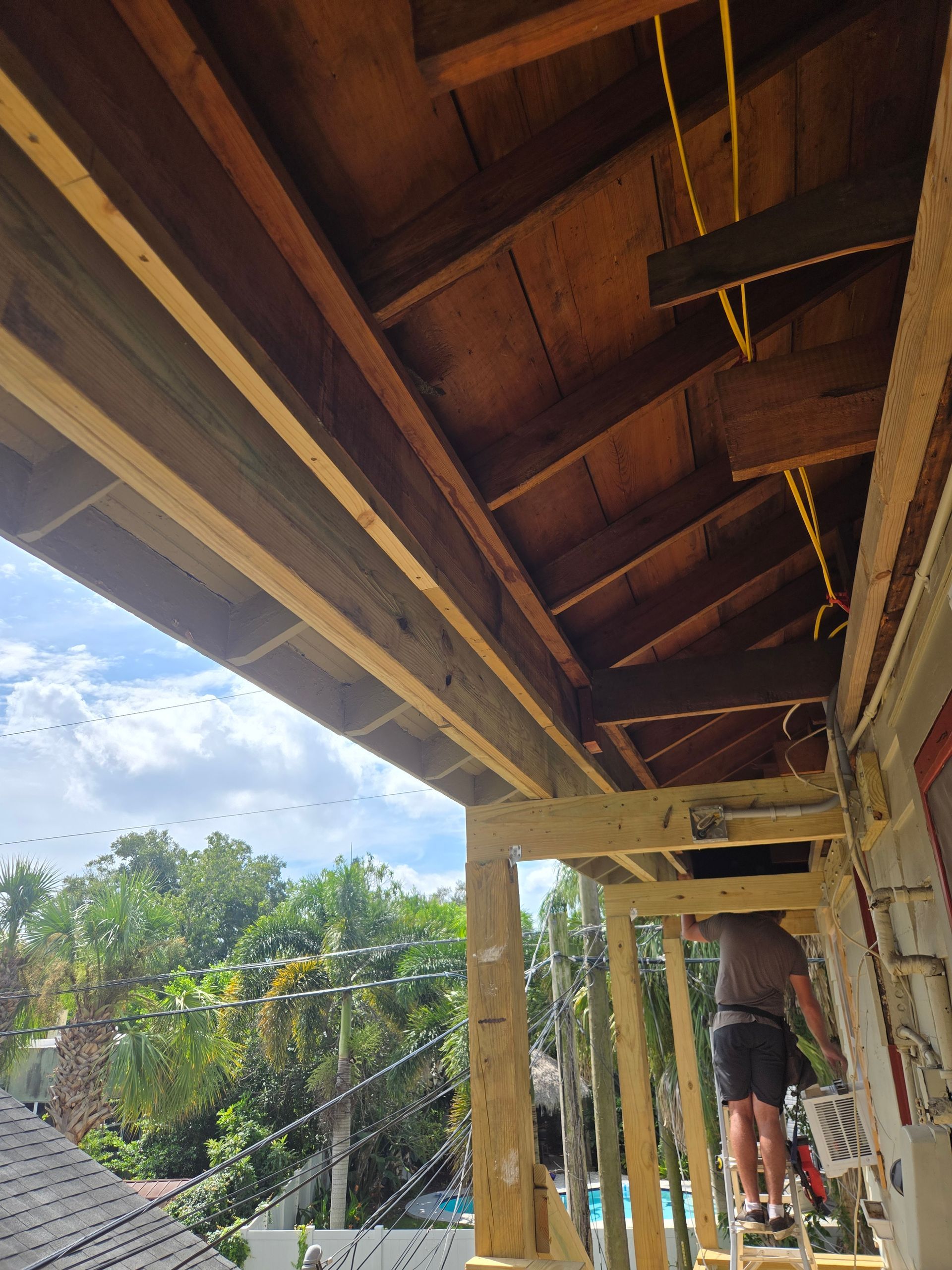 Construction worker on a ladder working on a wooden porch roof. Brown wood, yellow wires. Outdoors, bright.