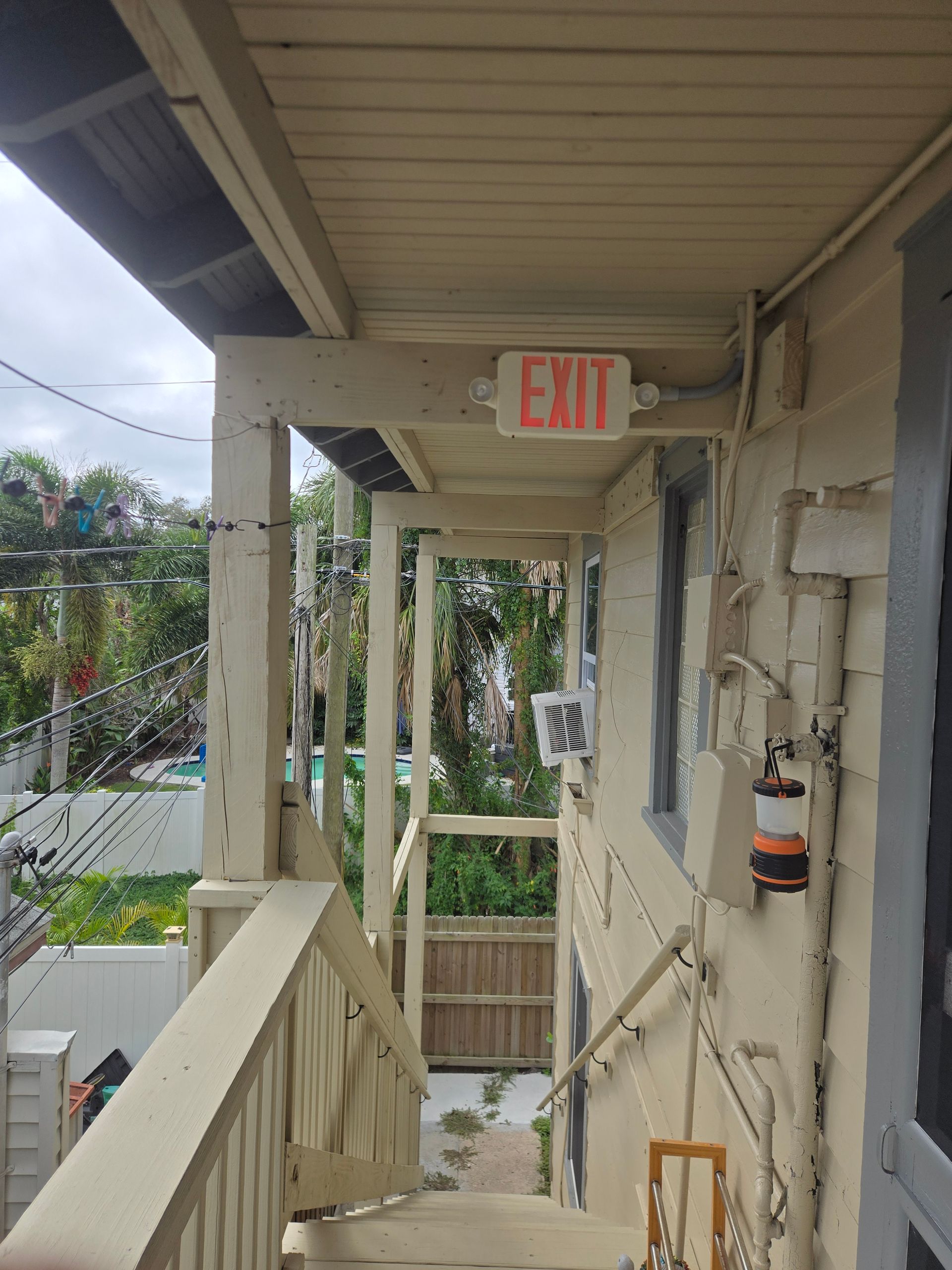 Staircase leading to exit sign on a porch with beige siding, railing, and a view of greenery.