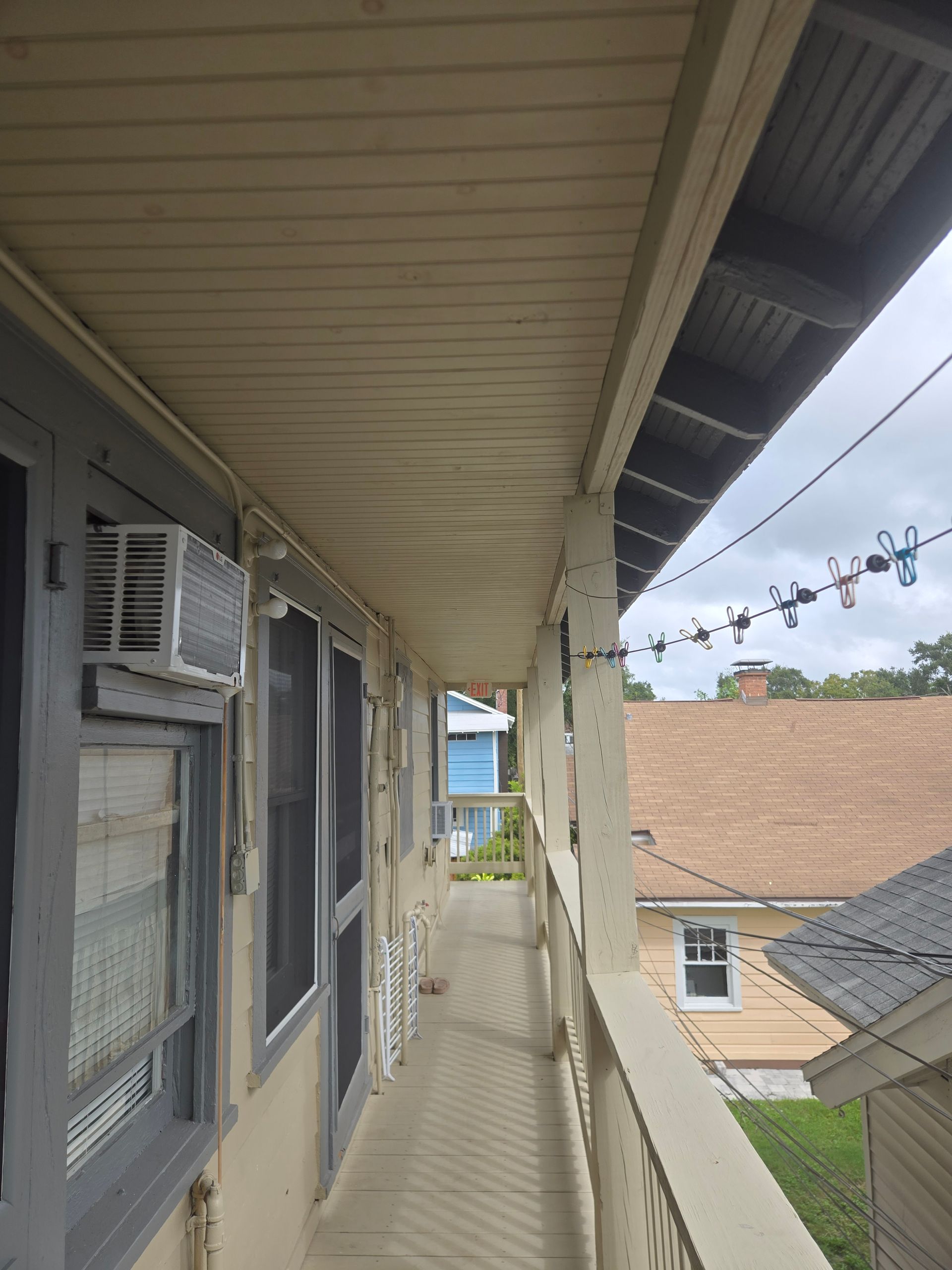 Narrow outdoor balcony with a light-colored ceiling and wooden railings, overlooking other buildings under a cloudy sky.