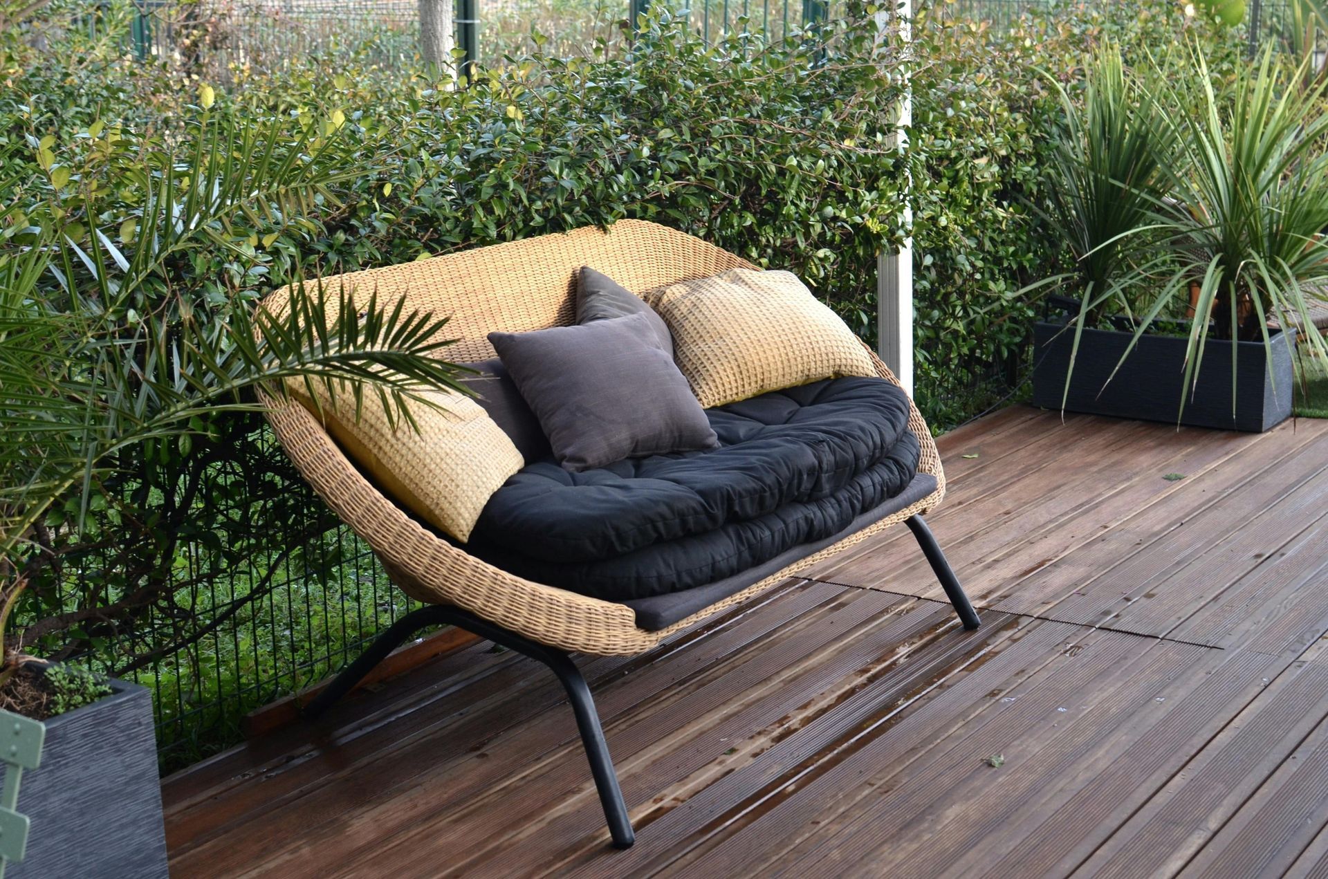 Woven outdoor loveseat with black cushions and pillows on a wooden deck. Green foliage in background.