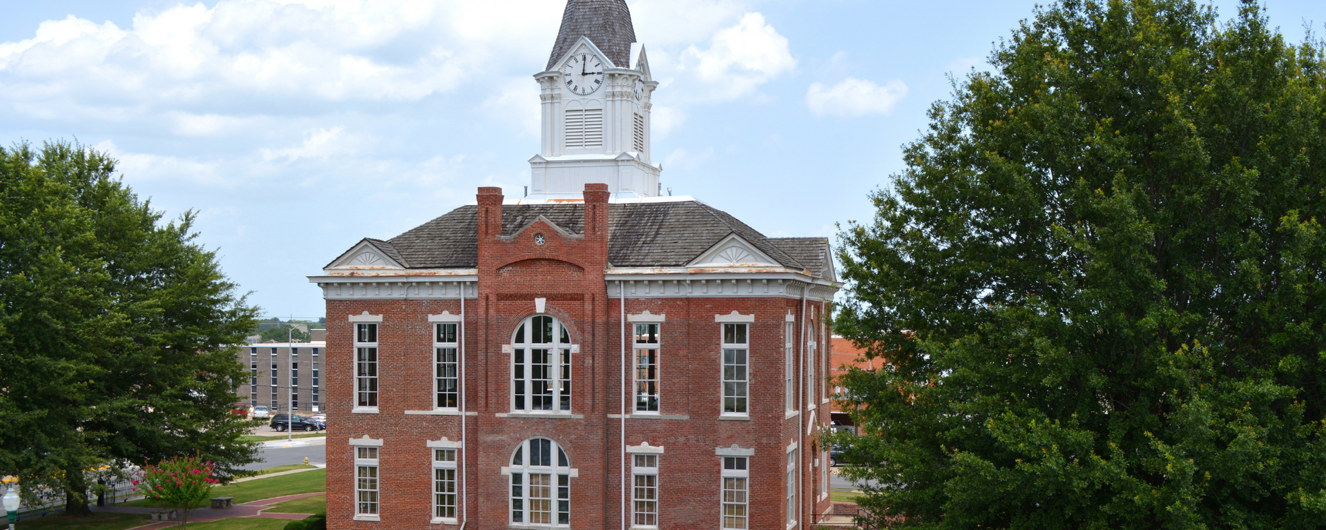 Red brick civic building with white clock tower, framed by green trees under a blue sky