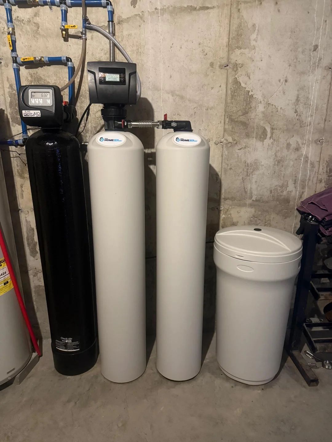 Water treatment system in a basement. Black tank, two white tanks, and a white salt tank. Blue pipes overhead.