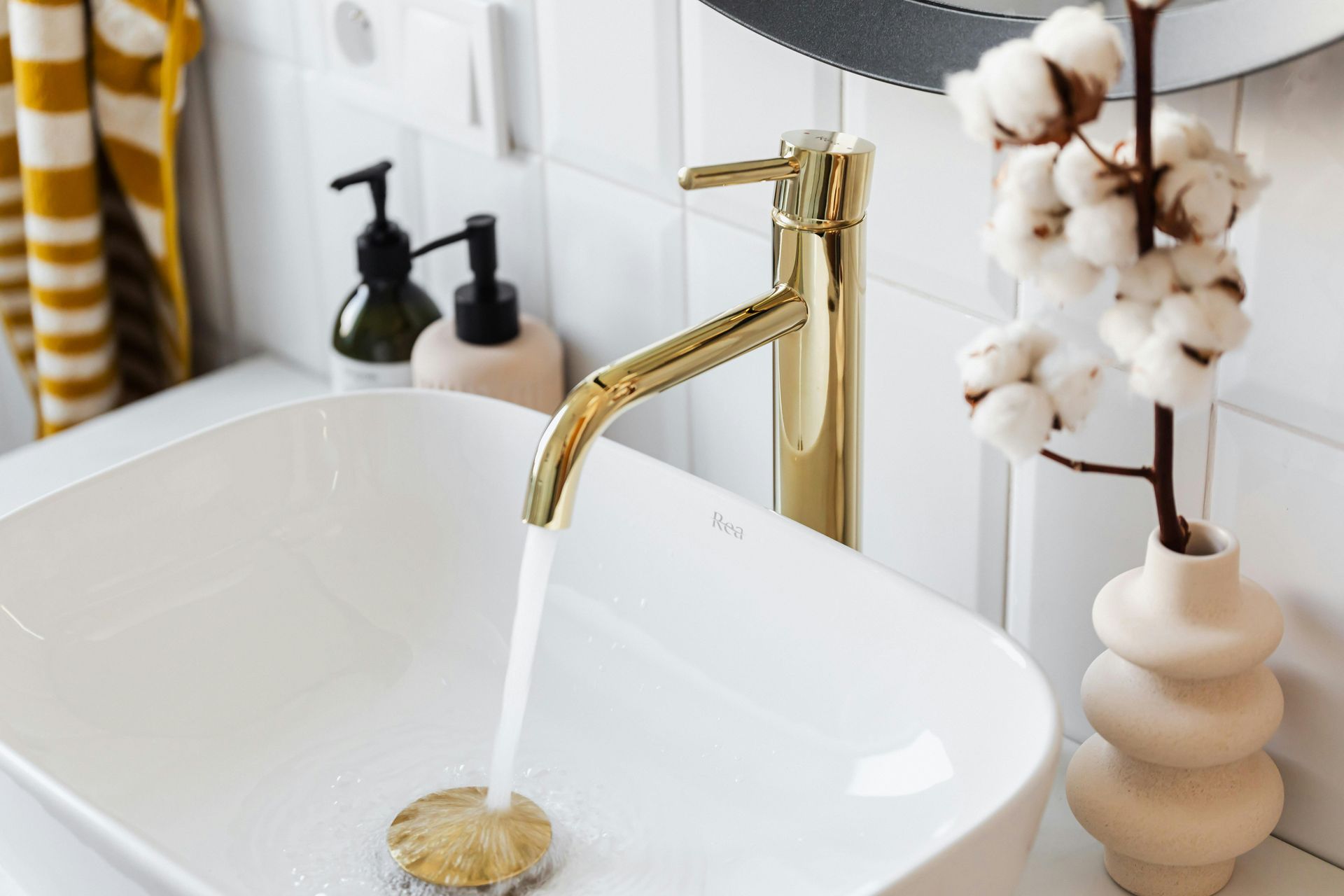 Gold faucet with running water into a white sink; cotton stems in a cream vase on the right.