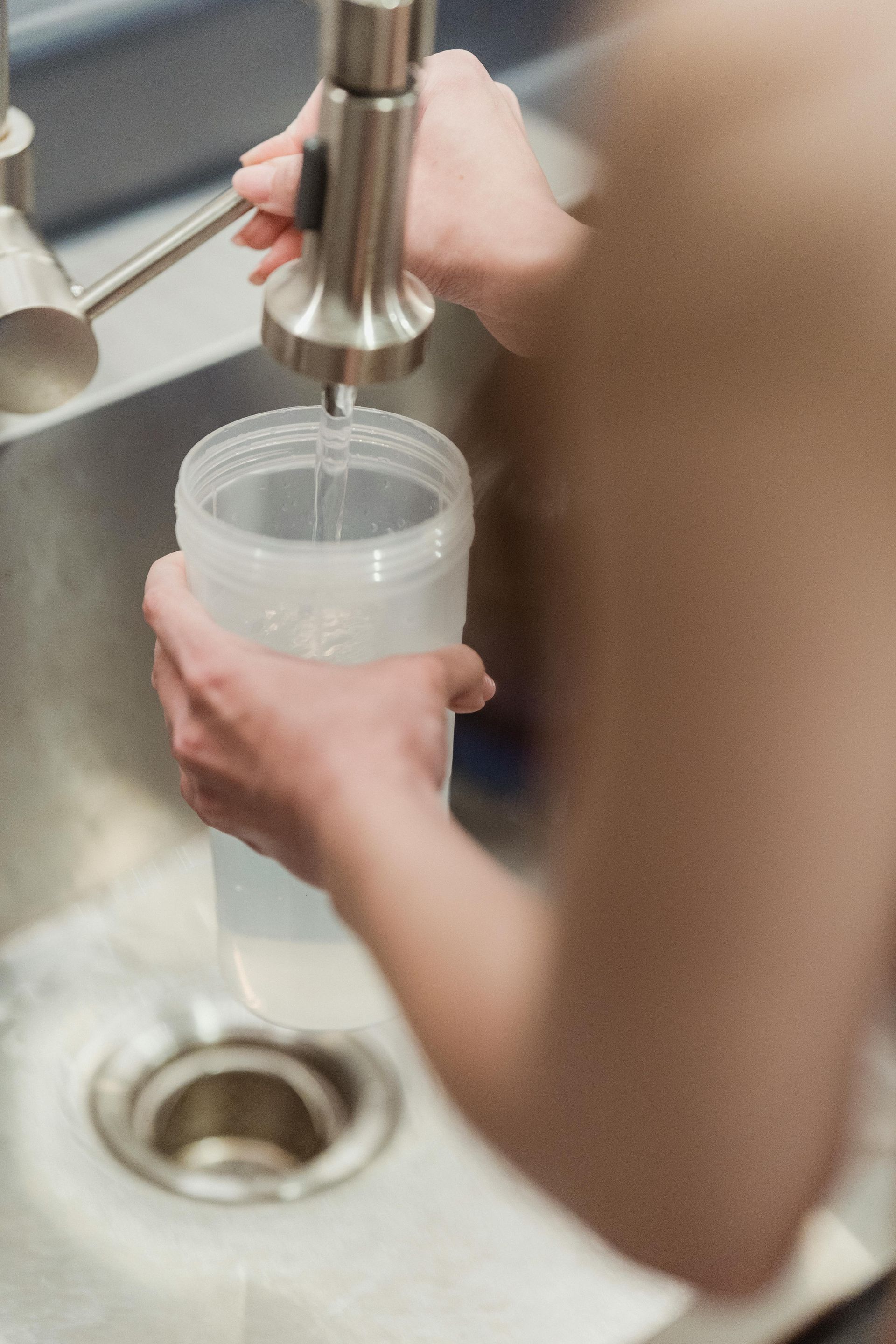 Person filling a tall, clear cup with water from a stainless steel faucet over a sink.