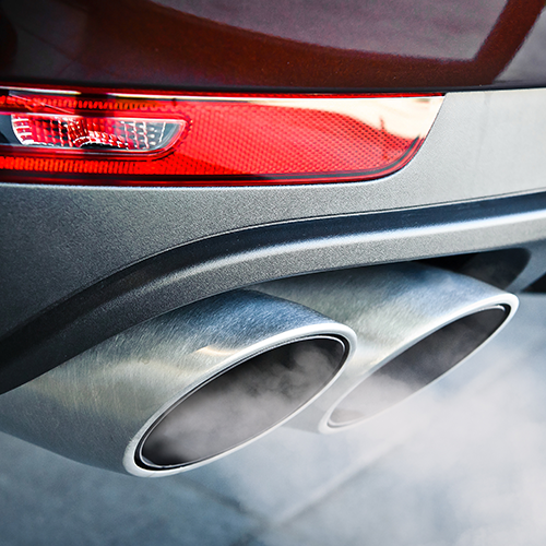 Close-up of a car's exhaust pipes with smoke coming out, near a red tail light.