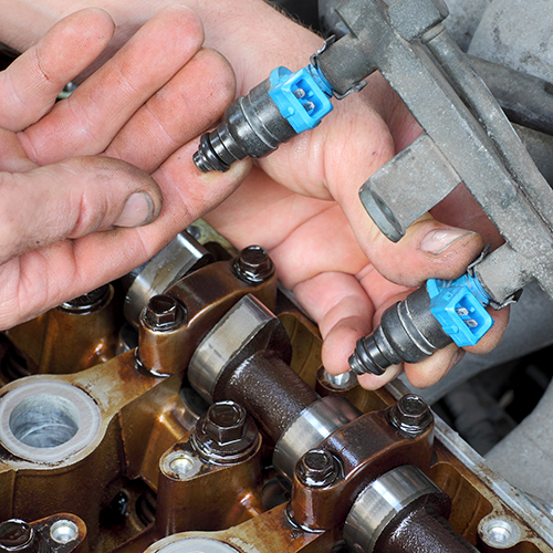 Hands installing fuel injectors on an engine with exposed camshafts and valve gear.