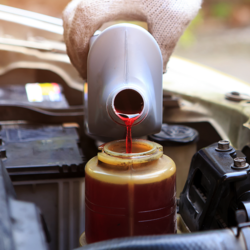 Gloved hand pouring red fluid from a container into a car reservoir.