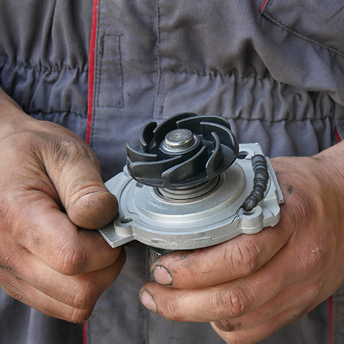 Hands holding a car water pump with a black impeller against a grey jumpsuit.