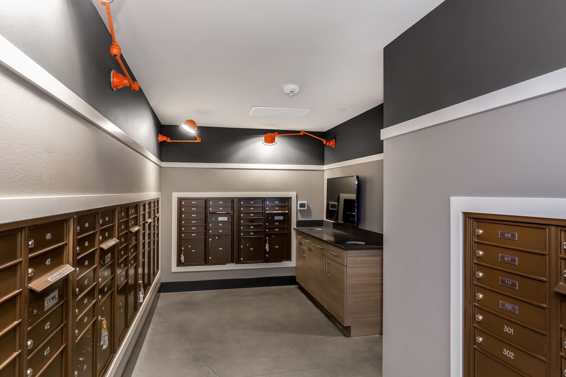Apartment mailboxes in a hallway, with a package drop off counter and a security camera, featuring brown, grey, and orange elements.