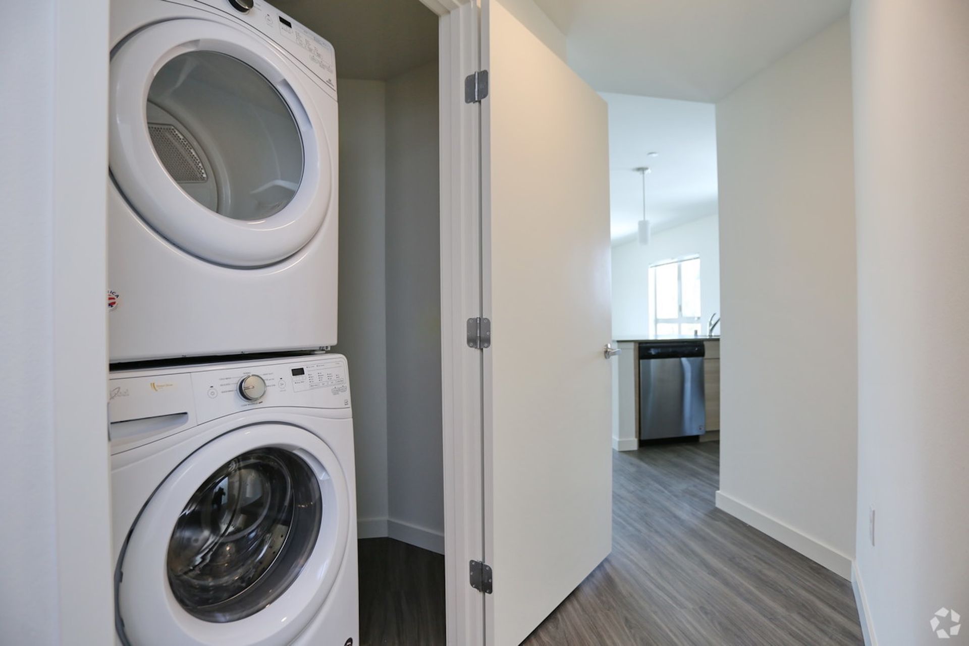 Stacked white washer and dryer in a laundry closet. Open door reveals hallway and kitchen.