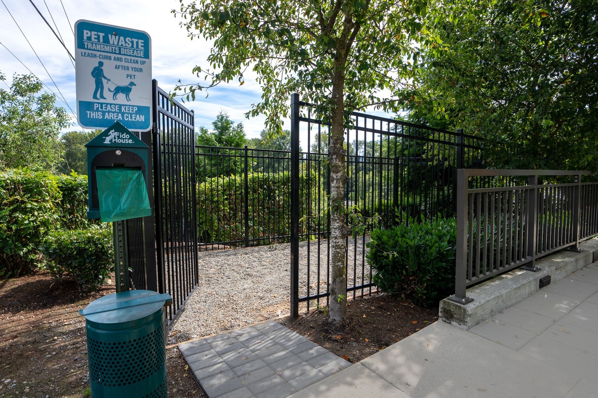 Dog waste station: sign, bag dispenser, and trash can within a fenced area; stone ground.