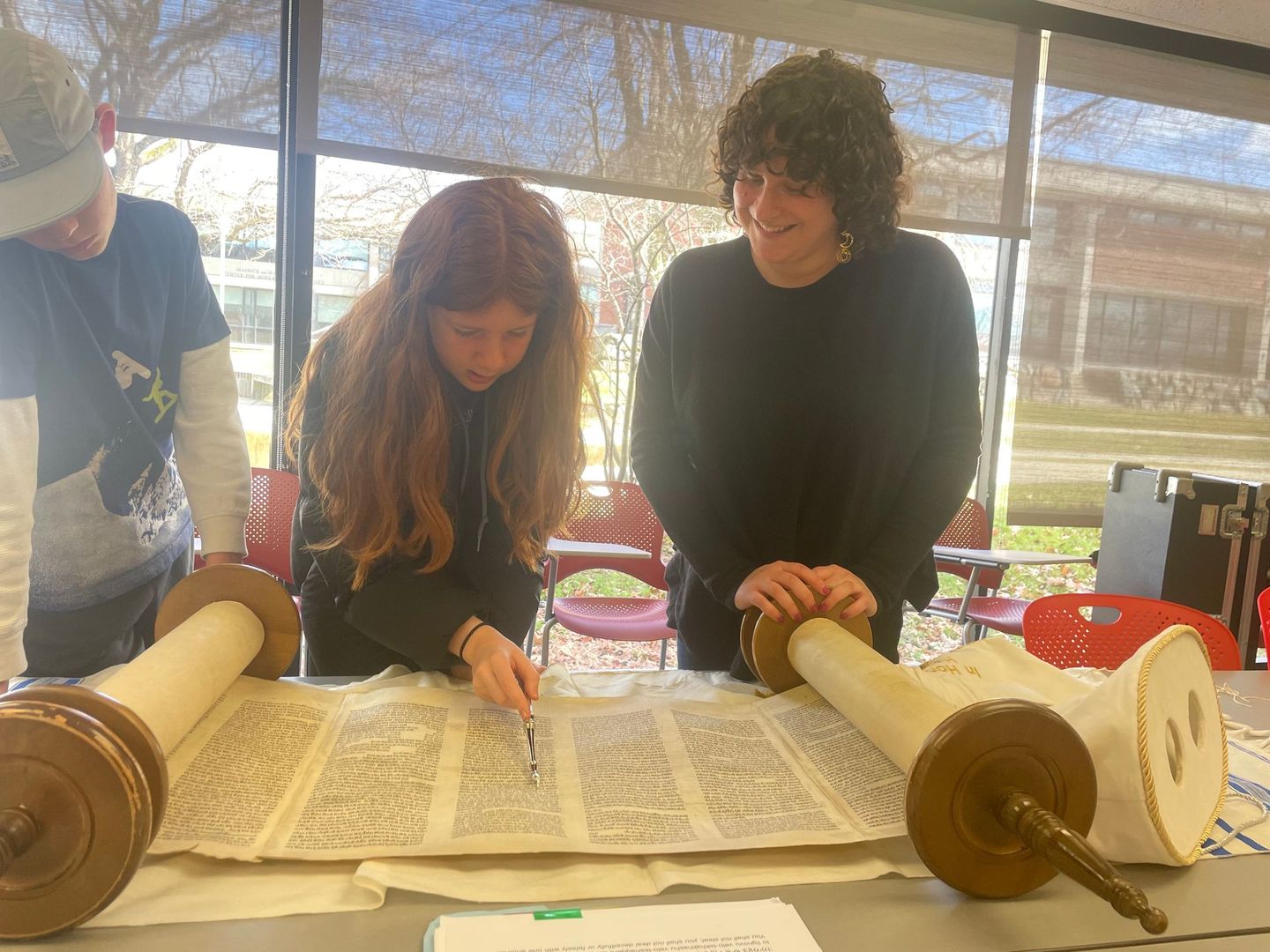 A group of people are sitting at a table reading a torah scroll.