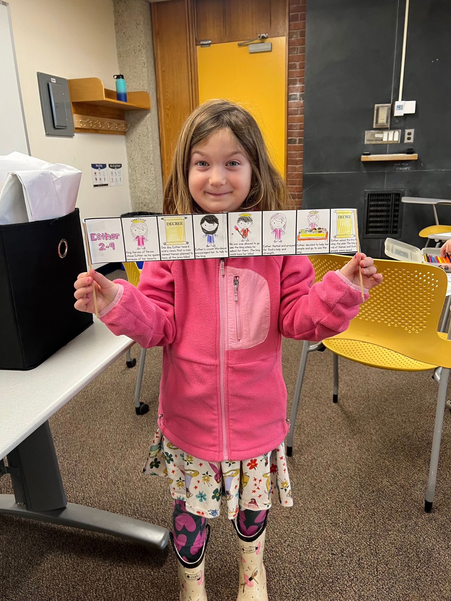 A little girl in a pink jacket is holding a sign in a classroom.