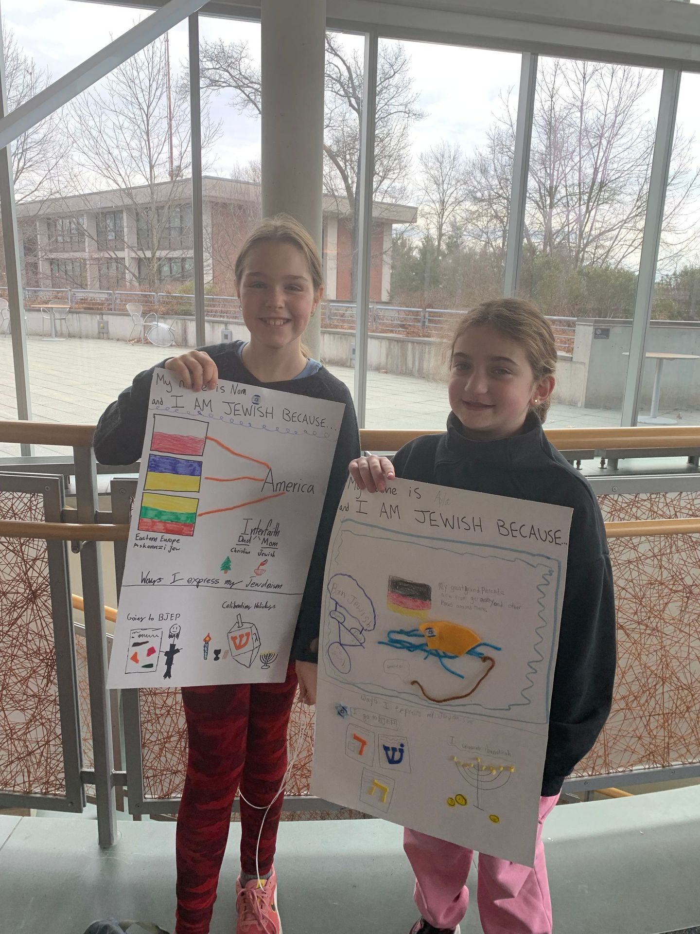 Two young girls are holding up their drawings in front of a window.