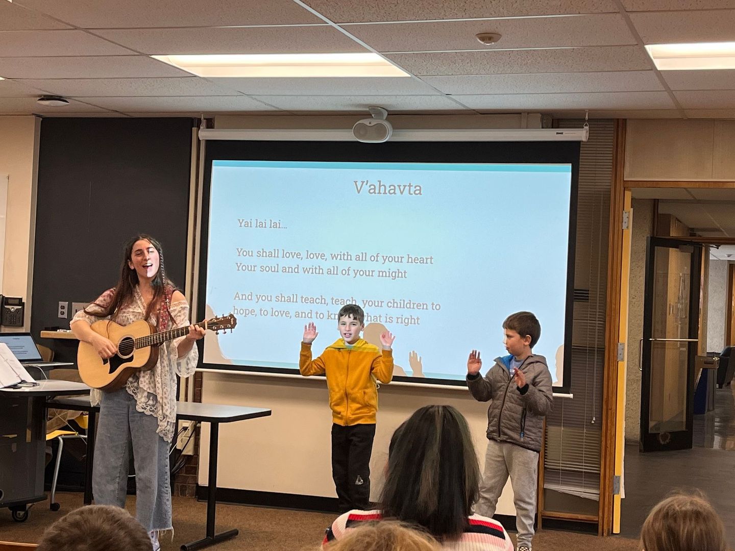 A woman is playing a guitar in front of a projector screen in a classroom.